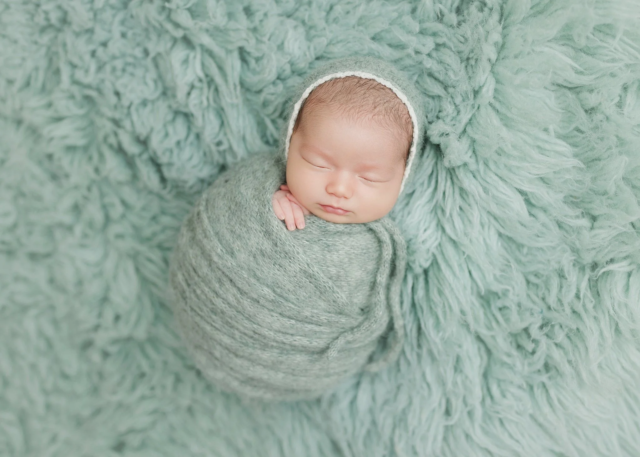 Sleeping newborn swaddled in a sage green knit wrap with a matching bonnet, resting on a fluffy green blanket with one tiny hand beside the cheek.