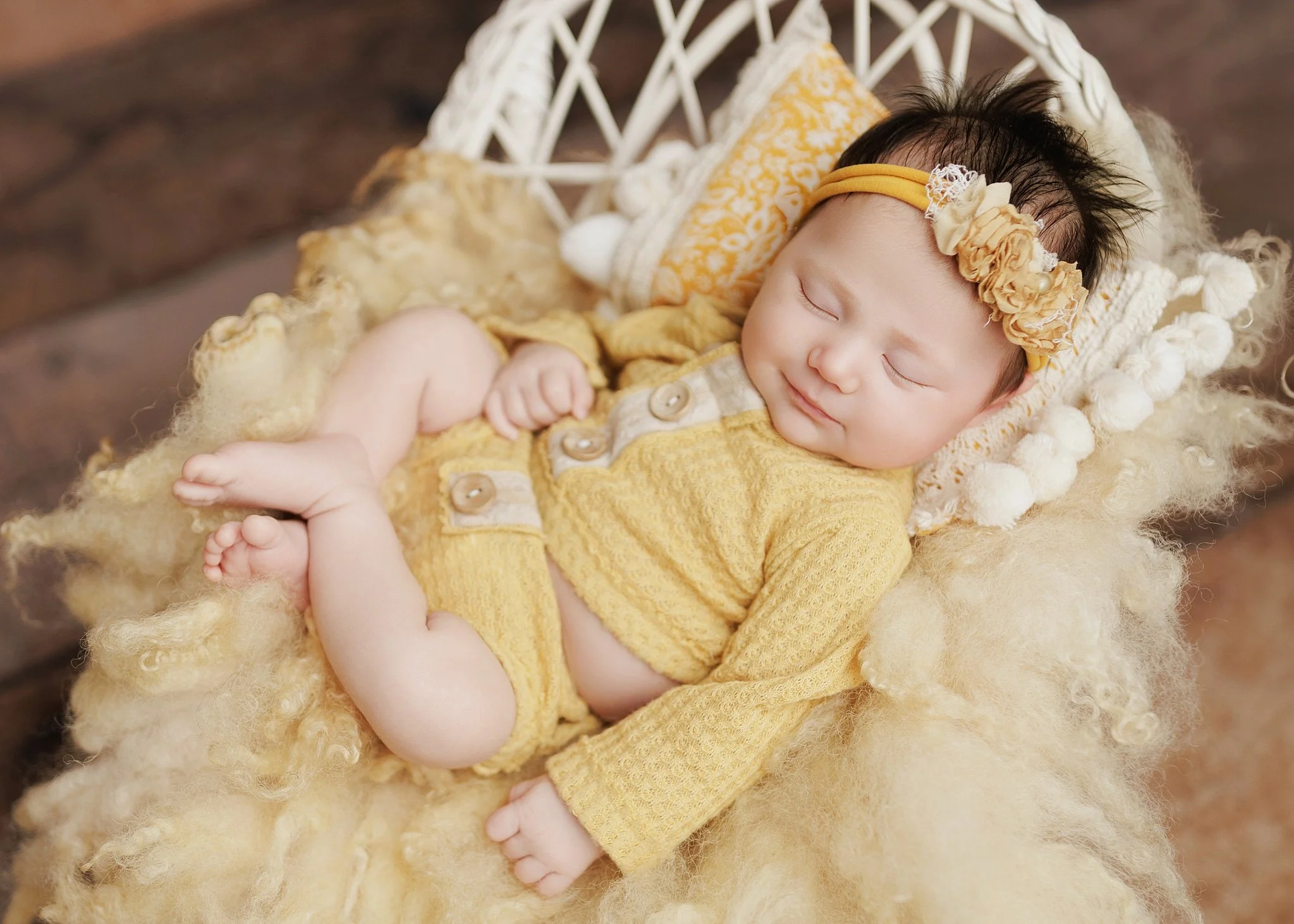 Sleeping newborn wearing a yellow knit outfit and matching floral headband, curled in a white wicker chair with yellow cushions and fluffy wool on a rustic wood backdrop.