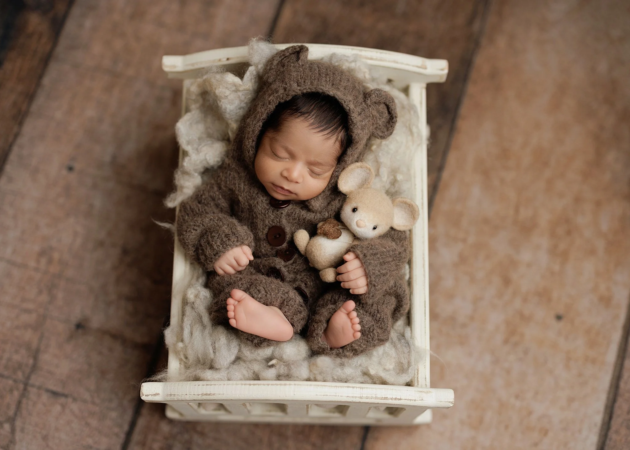Overhead portrait of a sleeping newborn boy in a brown bear-eared knit outfit, lying beside a tiny teddy bear in a miniature white wooden bed.