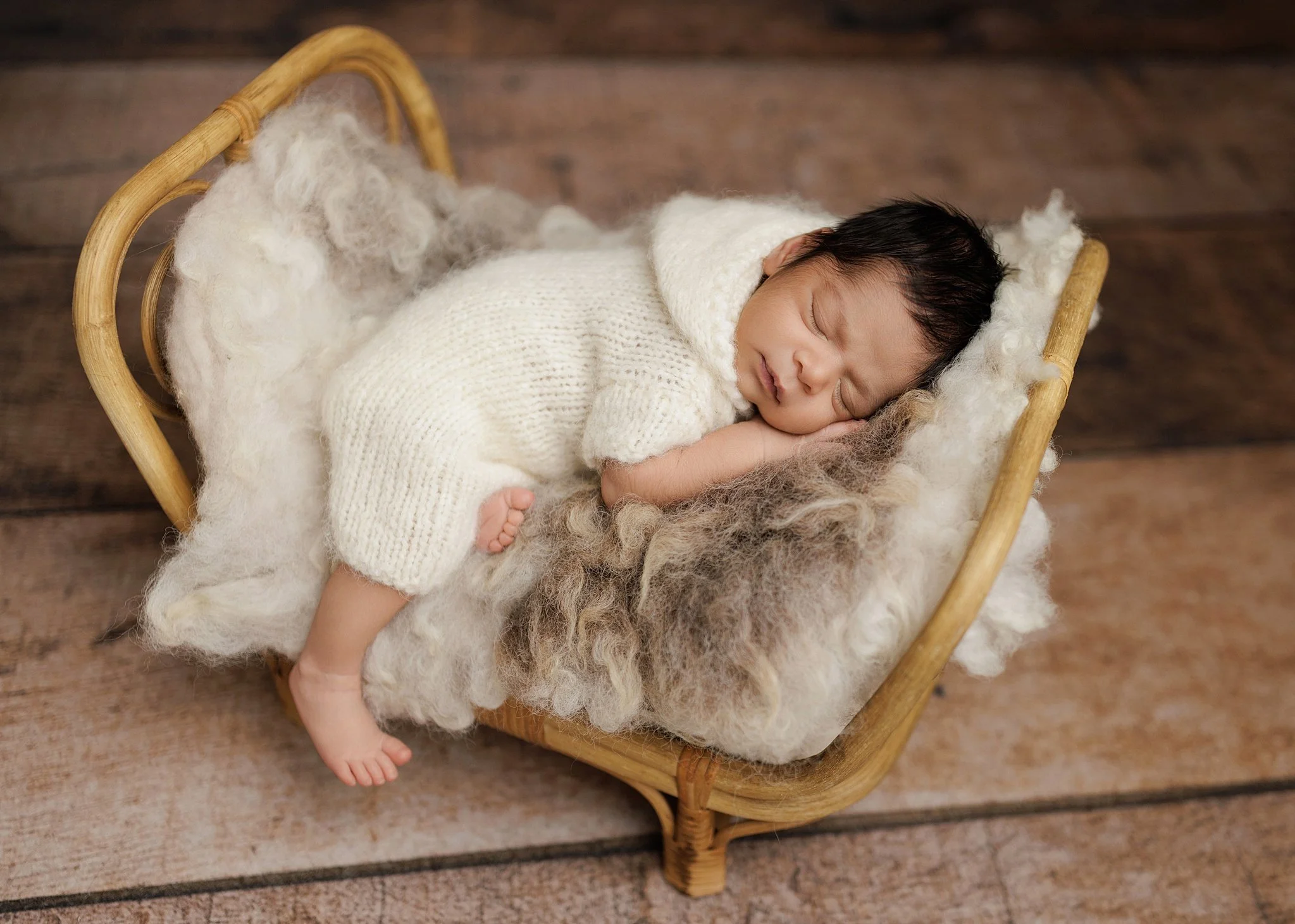 Sleeping newborn boy in a cream knit outfit, lying on his side in a small rattan bed with soft ivory and taupe layers against a wooden backdrop.
