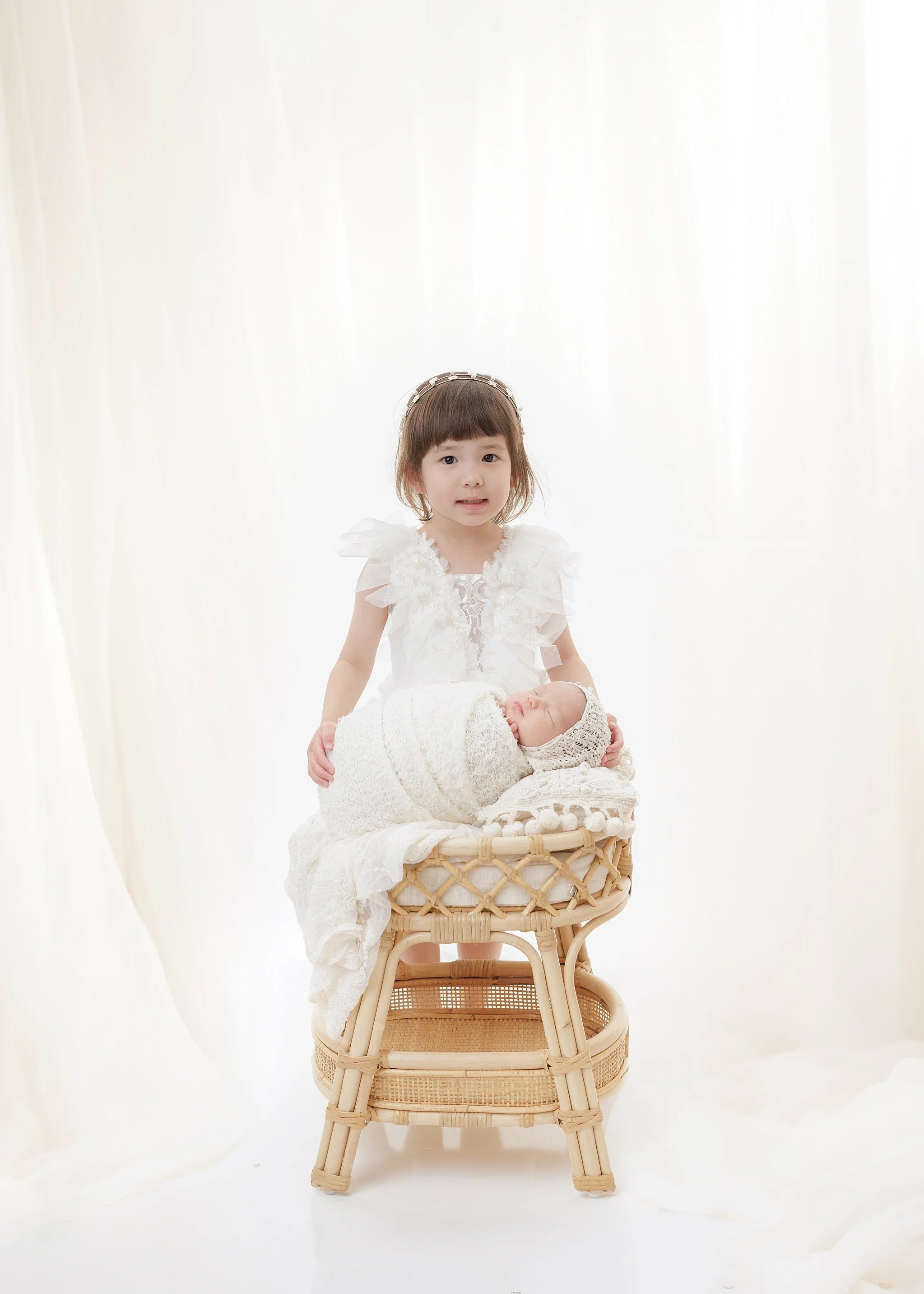 Big sister in a white dress standing behind a sleeping newborn wrapped in cream in a rattan bassinet, against soft white curtains.