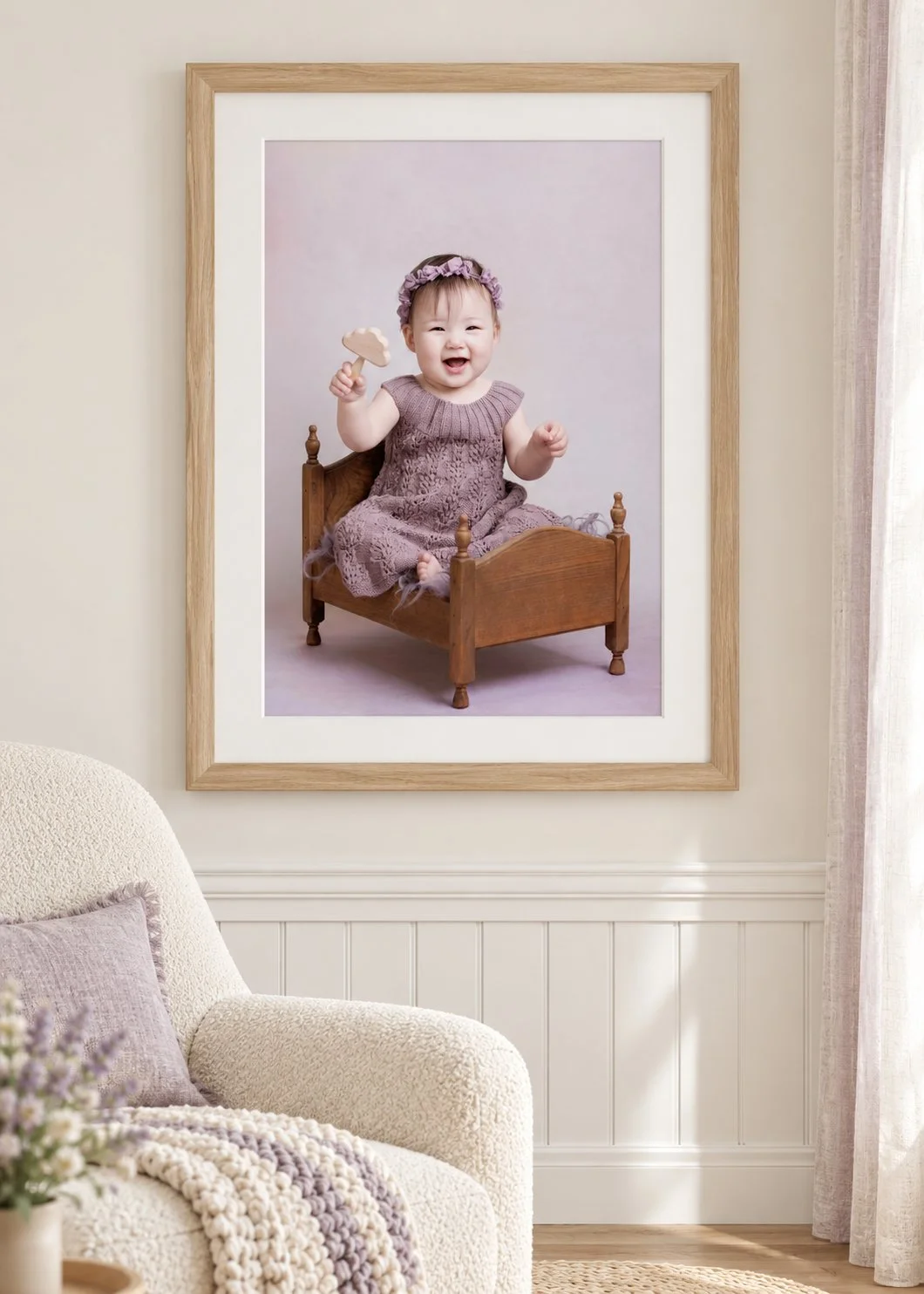 Baby portrait in a pale oak frame with a white mat above a cream nursery chair, featuring a smiling baby girl in a lavender dress holding a wooden toy.