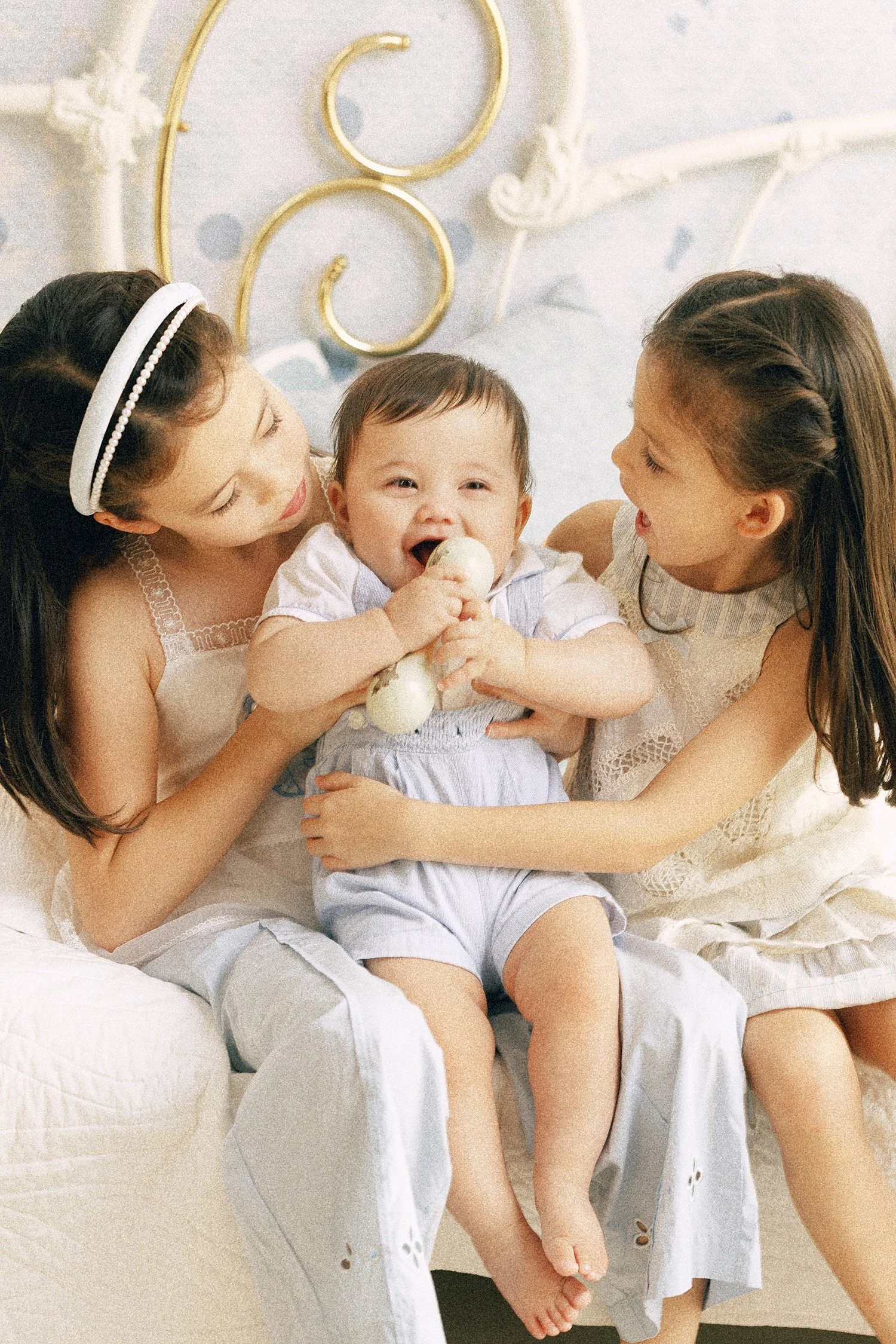 Two sisters cuddling their baby brother on a vintage bed during a Storybook Bedroom sibling portrait session in Southlake, Texas.