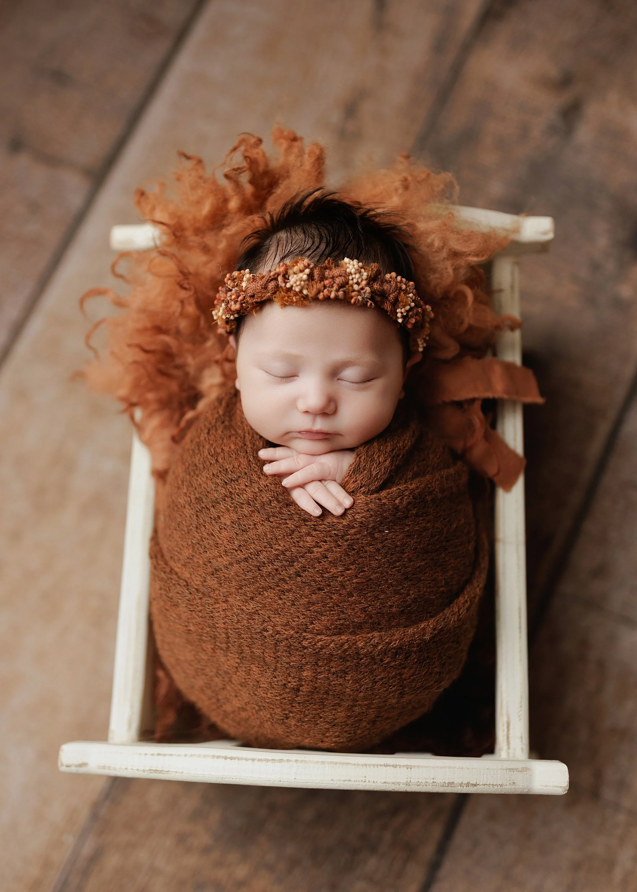 Overhead portrait of a sleeping newborn in a rust-colored wrap and autumn floral headband, with hands beneath the chin in a miniature white wooden bed.