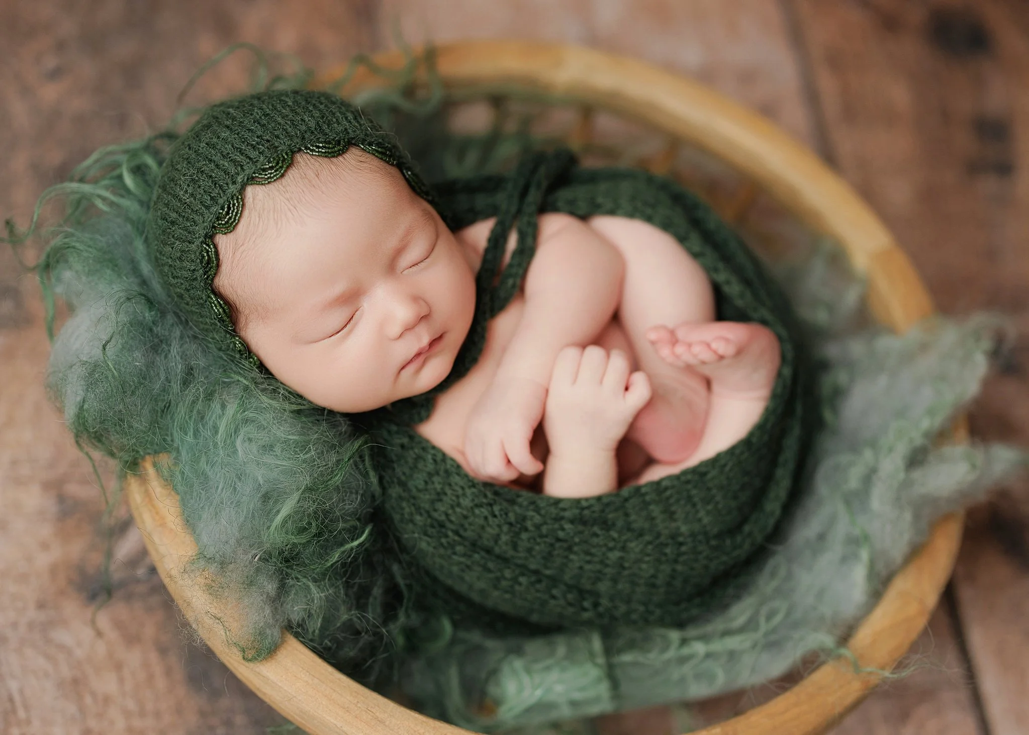 Close-up of a sleeping newborn wearing a forest green knit bonnet and matching wrap, curled in a wicker basket with tiny hands and feet visible.