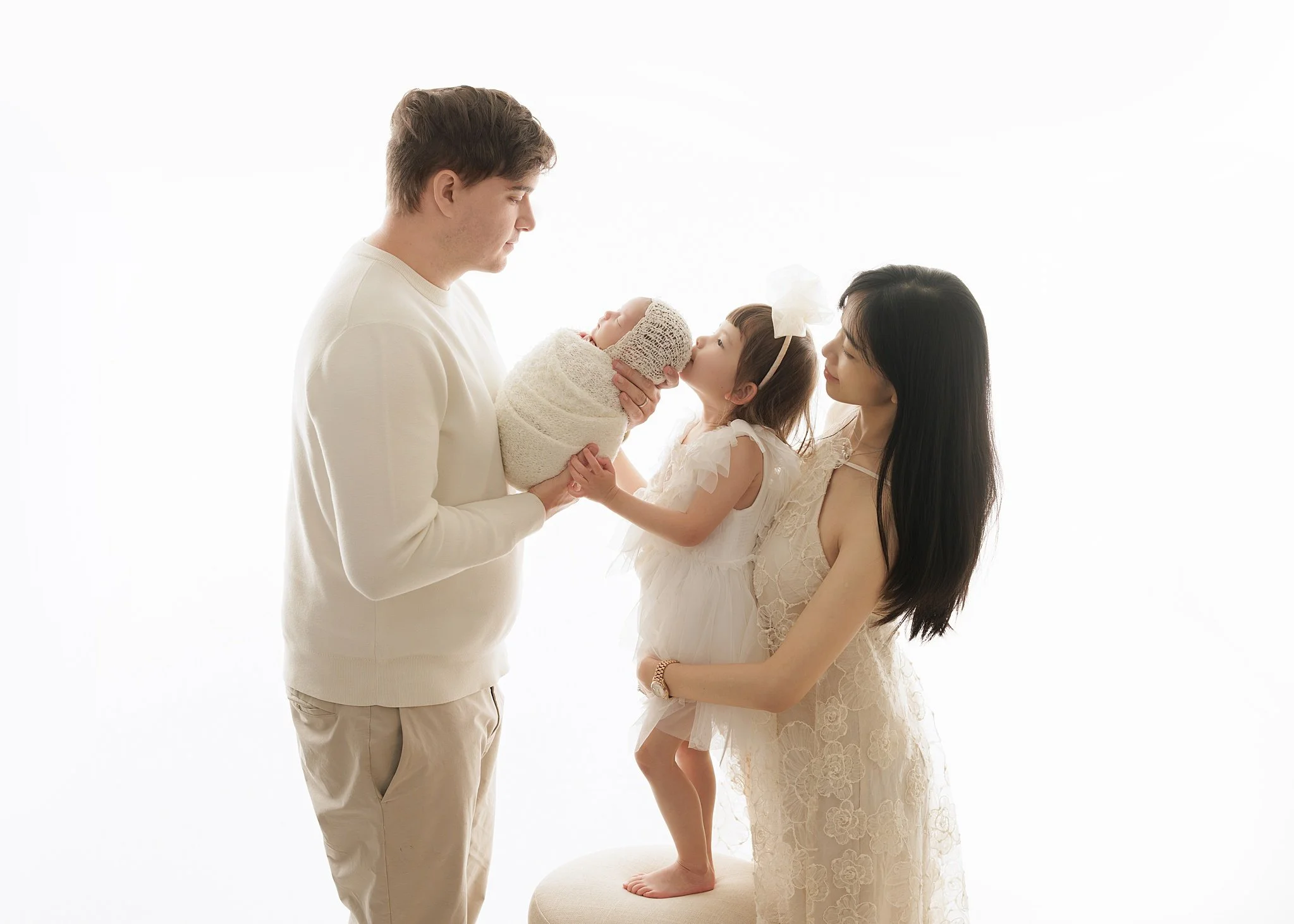 Father holding a newborn wrapped in cream as big sister kisses the baby’s head and mother supports her, against a bright white studio background.