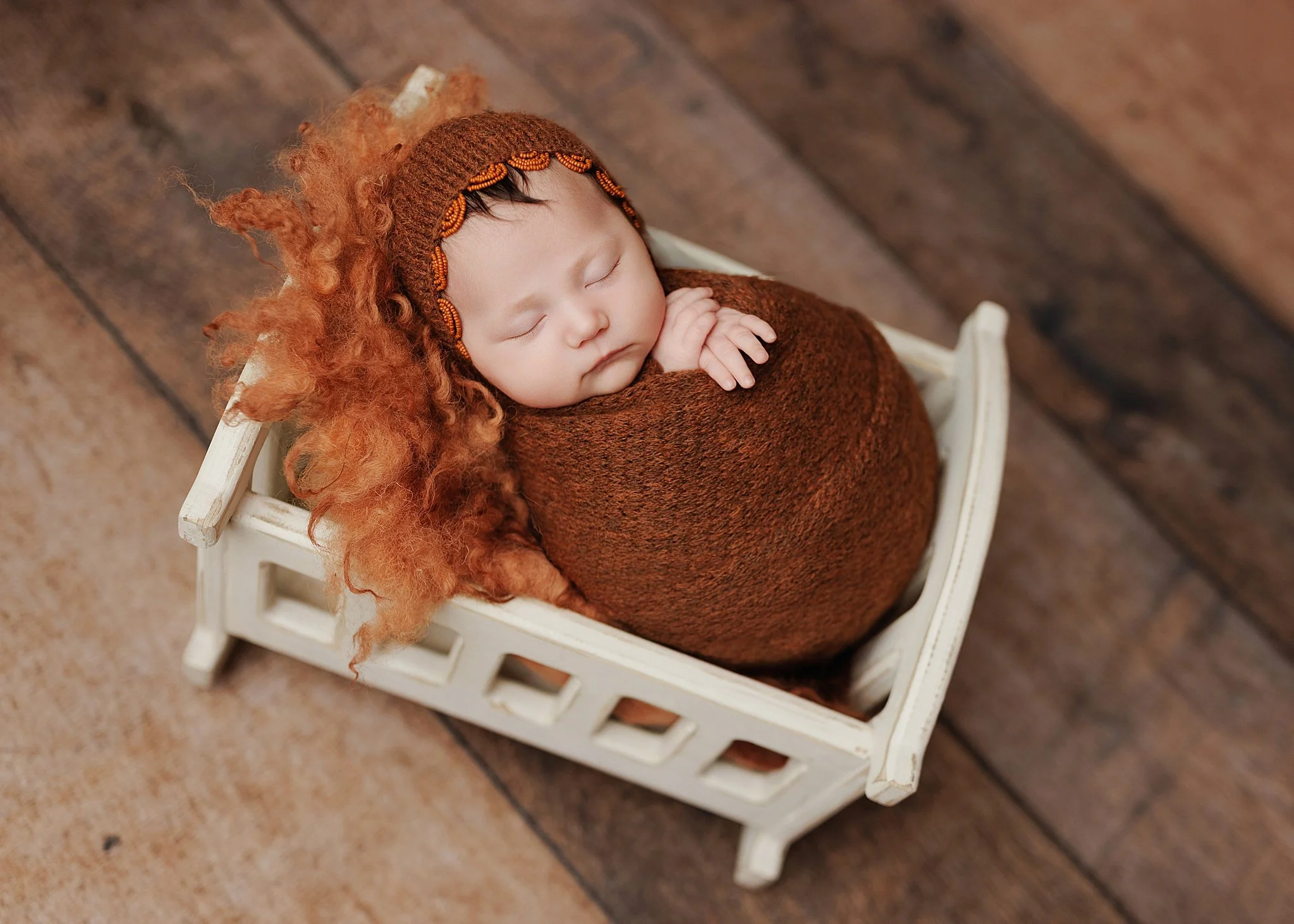 Sleeping newborn swaddled in a rust-colored wrap and matching knit bonnet, resting in a miniature white wooden bed with orange wool on a rustic wood backdrop.