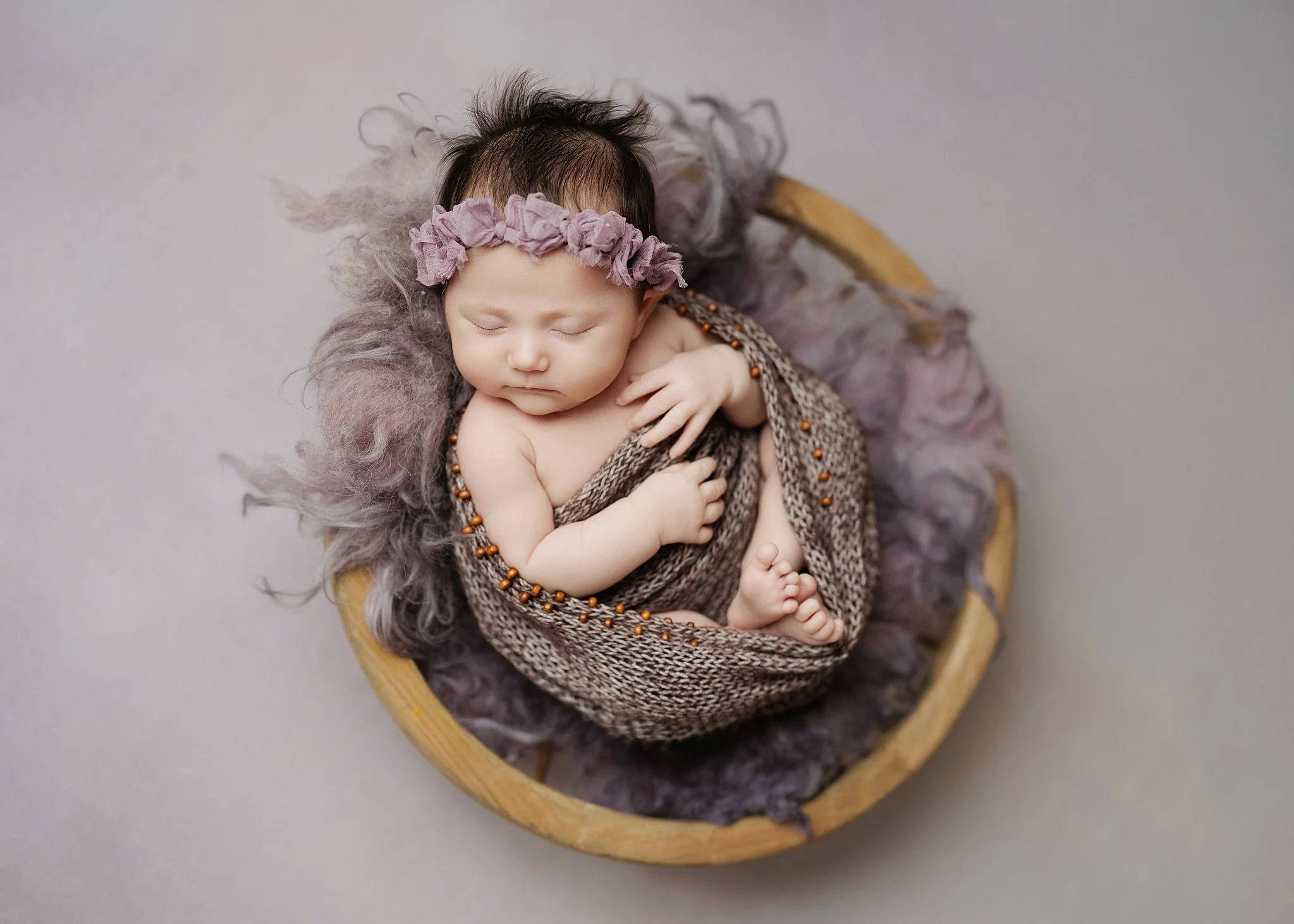 Sleeping newborn wearing a mauve floral headband and brown beaded knit wrap, curled in a wooden bowl with soft purple wool and tiny toes peeking out.