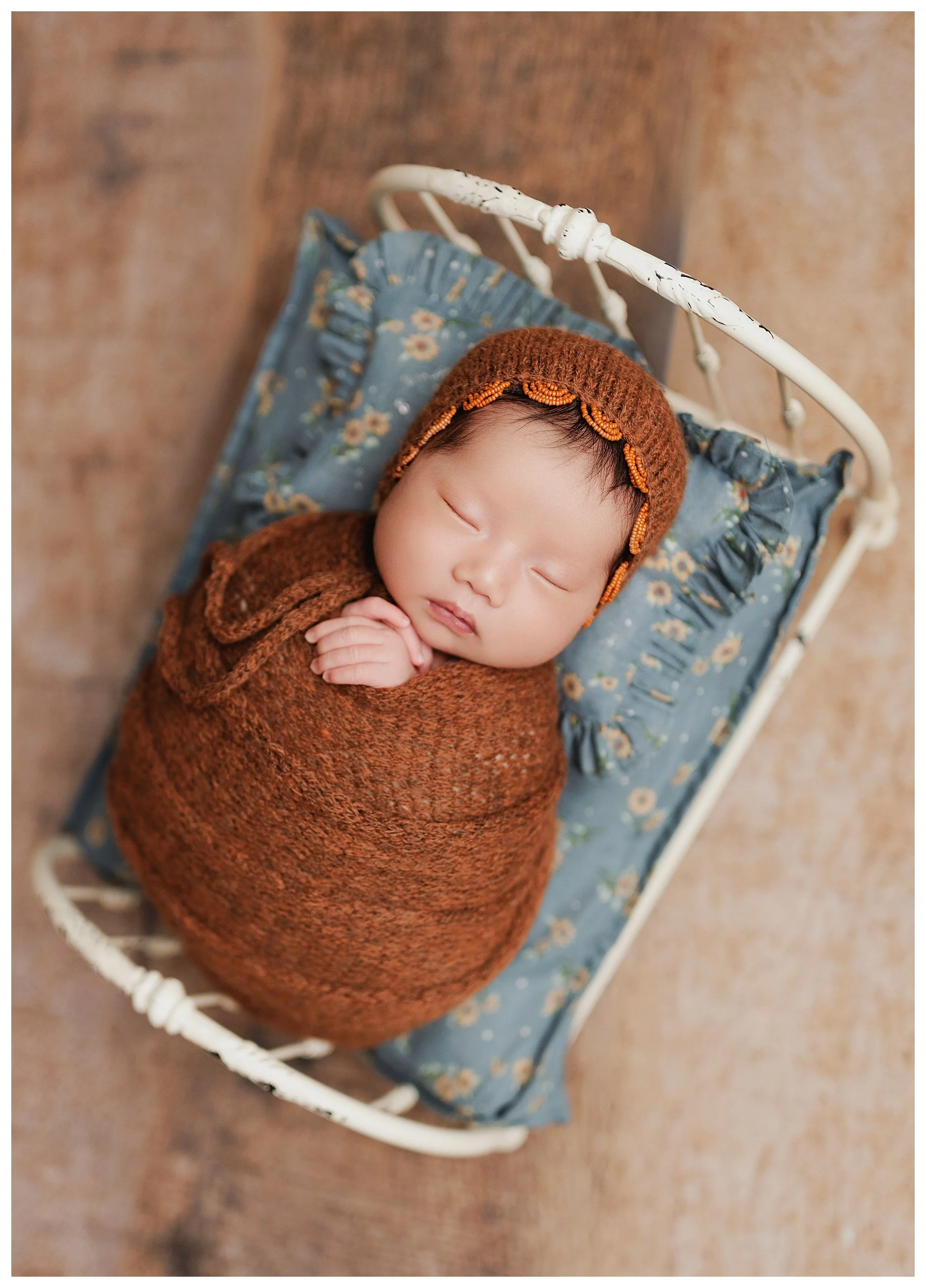 Sleeping baby girl wrapped in warm rust tones on a blue floral pillow during a Dallas newborn photo session