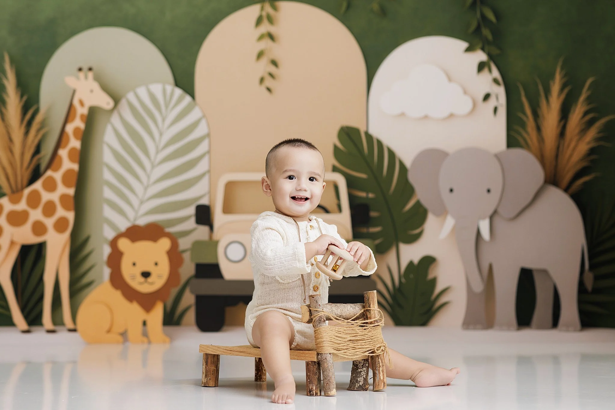 Smiling baby boy in a cream outfit sitting among giraffe, lion, and elephant decorations during a jungle-themed first birthday session.
