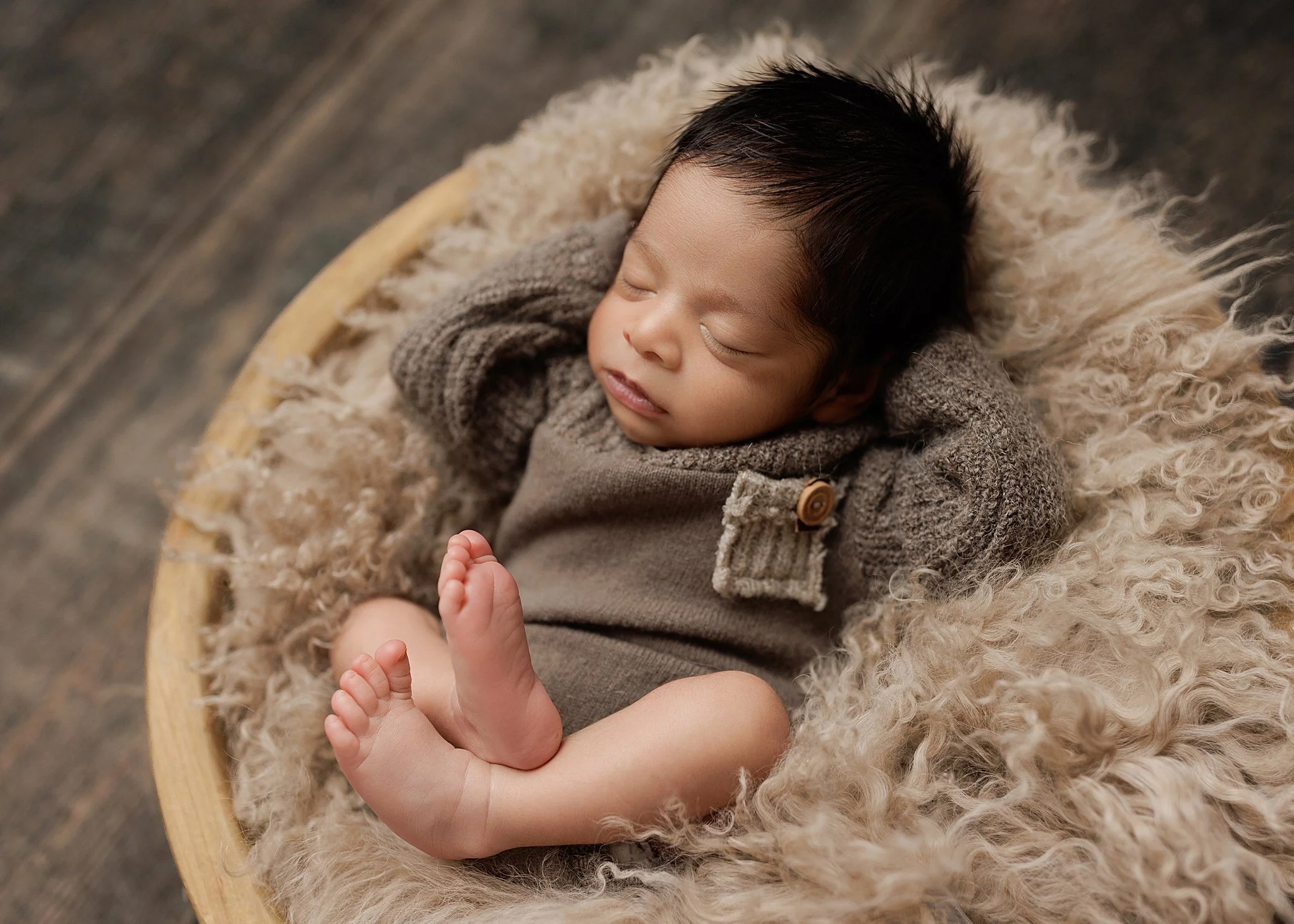 Sleeping newborn boy with dark hair wearing a taupe knit outfit, curled in a wooden bowl with beige textured layers and bare feet visible.