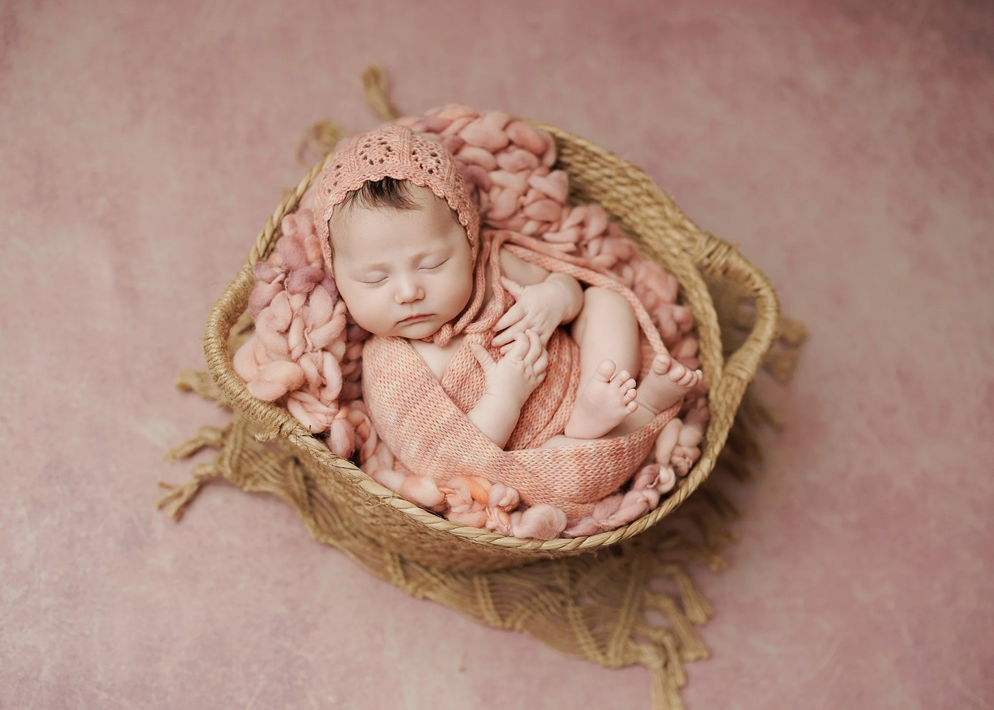 Sleeping newborn in a blush pink knit bonnet and matching wrap, curled in a woven basket lined with chunky pink wool against a soft pink backdrop.