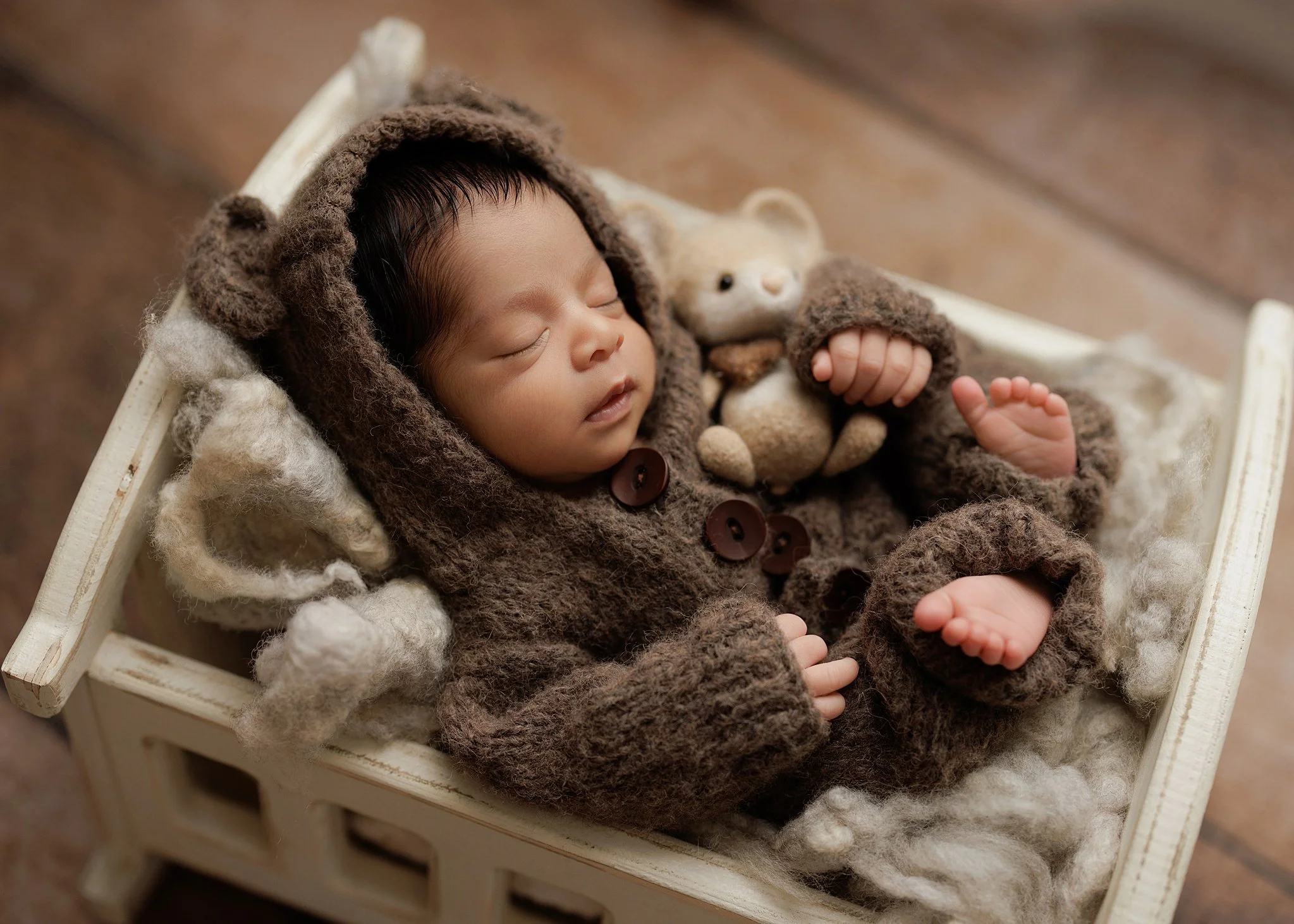 Close-up of a sleeping newborn boy in a brown hooded knit outfit, cuddling a tiny teddy bear with bare toes visible.