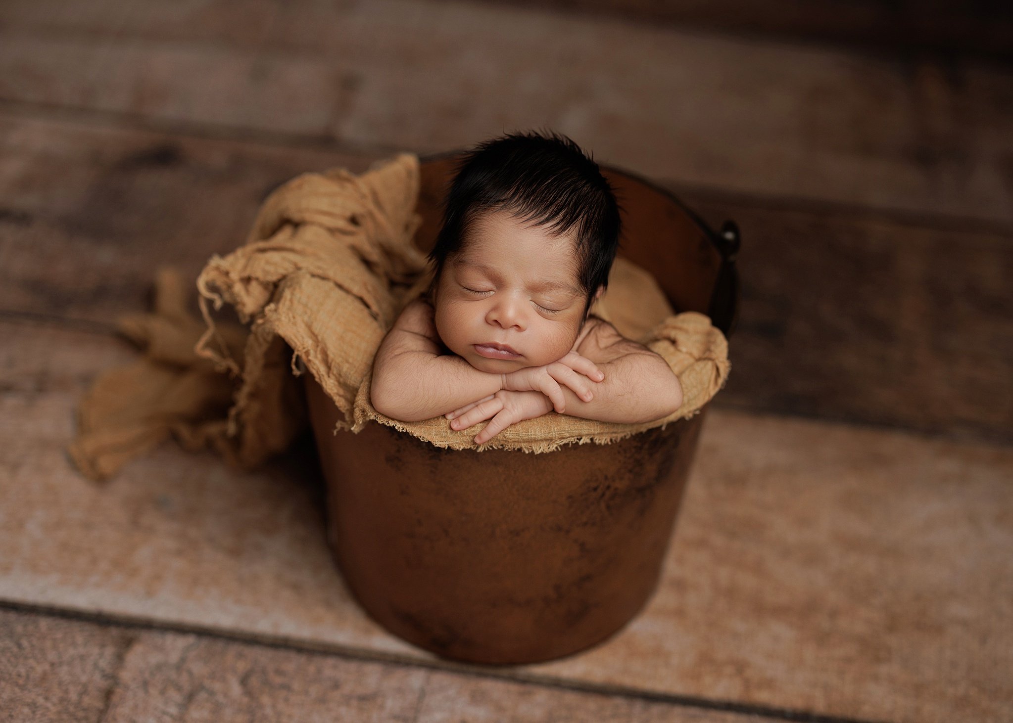 Sleeping newborn boy resting his chin on folded hands in a rustic brown bucket lined with caramel fabric, against a wooden backdrop.