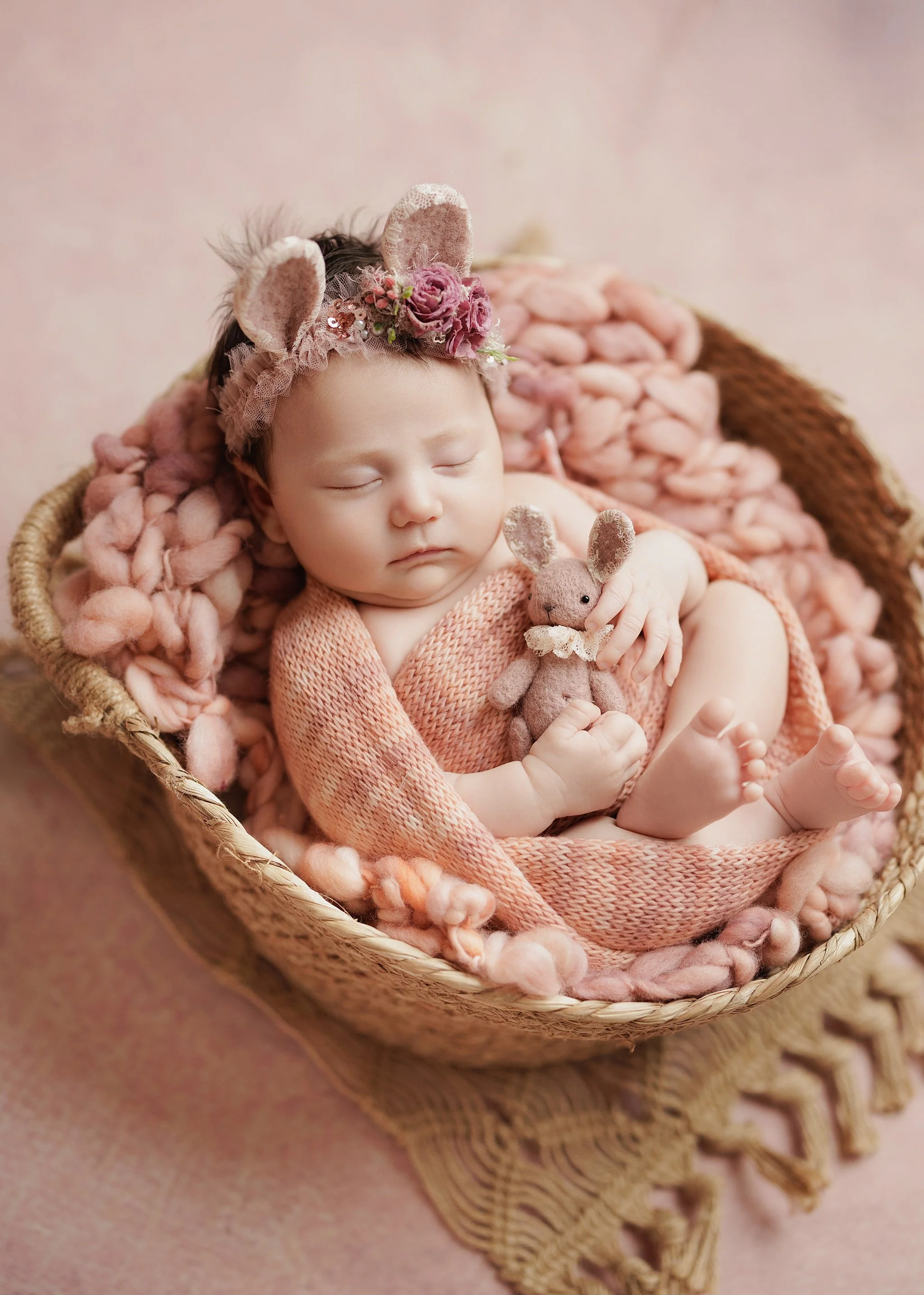 Sleeping newborn wearing a floral bunny-ear headband and pink knit wrap, holding a tiny stuffed bunny in a woven basket with soft pink wool.