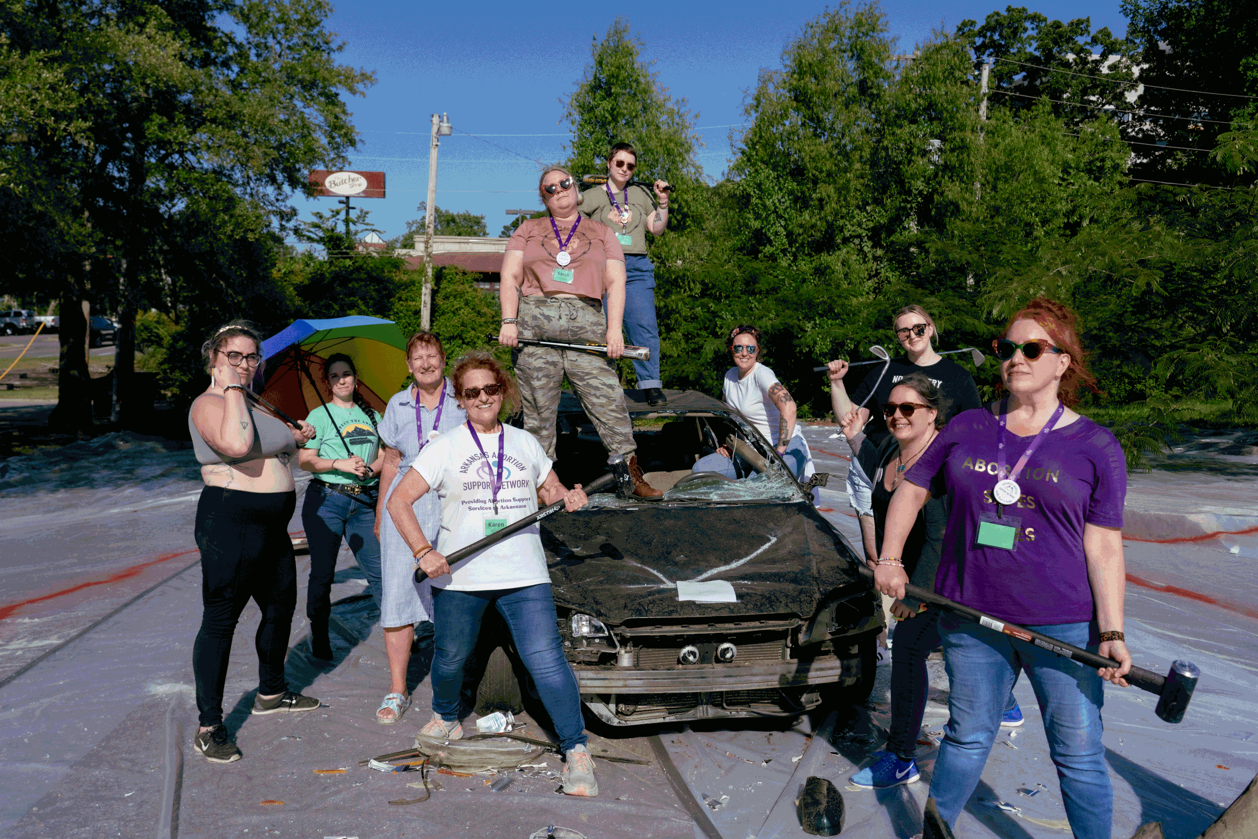 A group of femme-presenting people strike power poses on and around a smashed up car. Several weild sledgehammers, baseball bats, or golf clubs. They look like they're on the cover of a very cool album.
