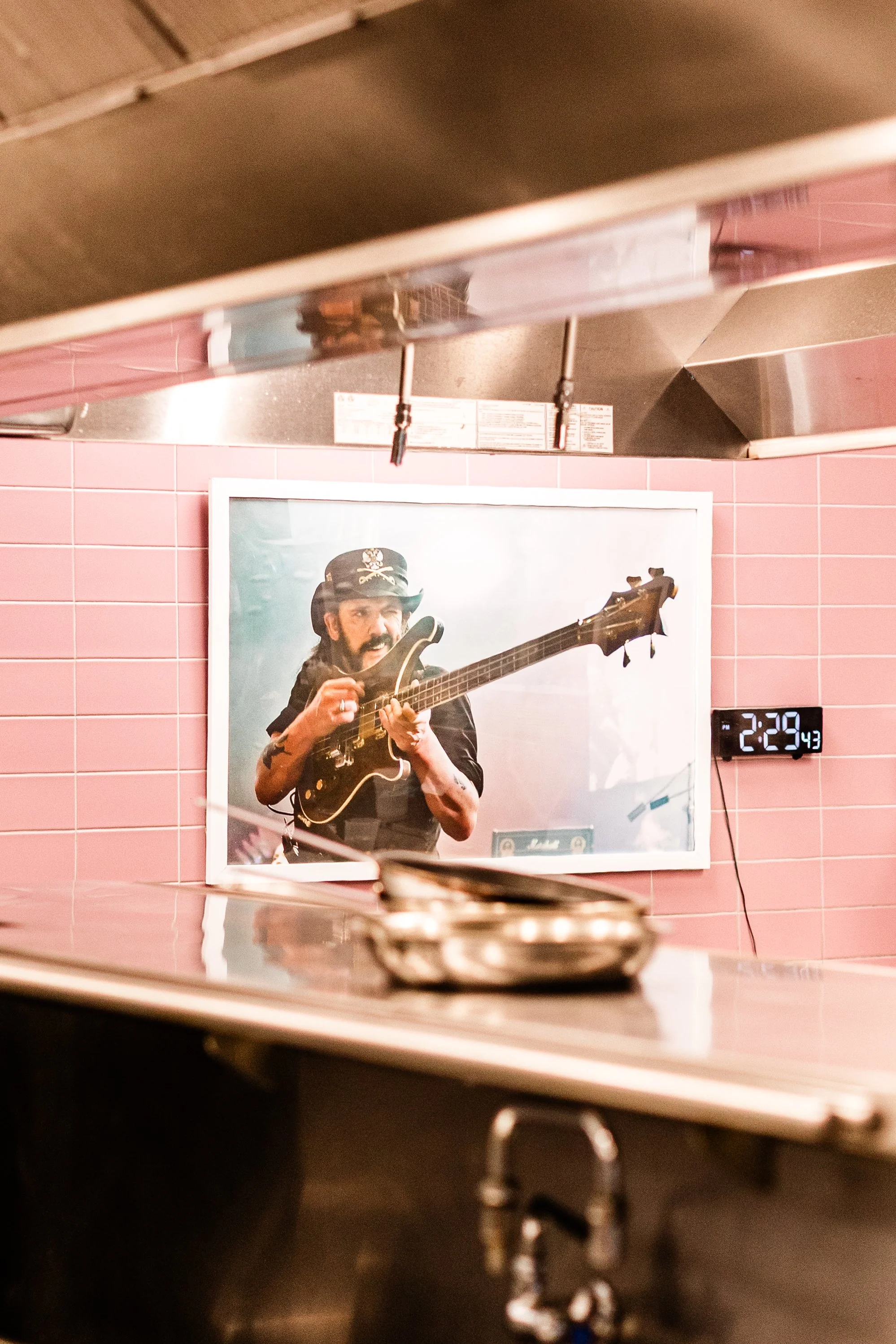 A person wearing a black hat with a skull and crossbones design playing an electric guitar, reflected in a mirror on a pink tiled wall, with a digital clock showing 2:29 PM in the background.
