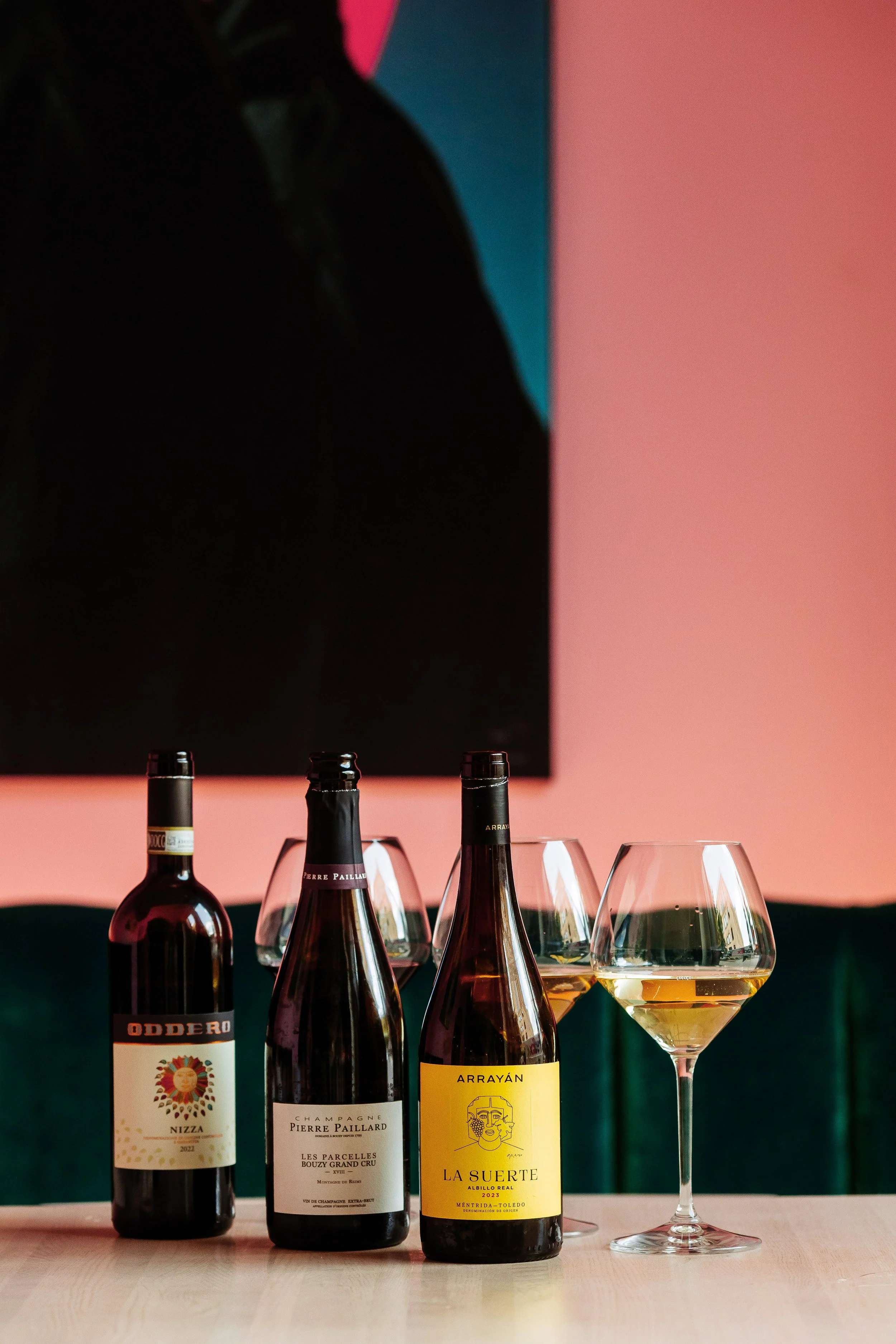 Five wine bottles and three wine glasses with white wine on a light wood table, with a pink and teal background and a black rectangle mounted on the wall.