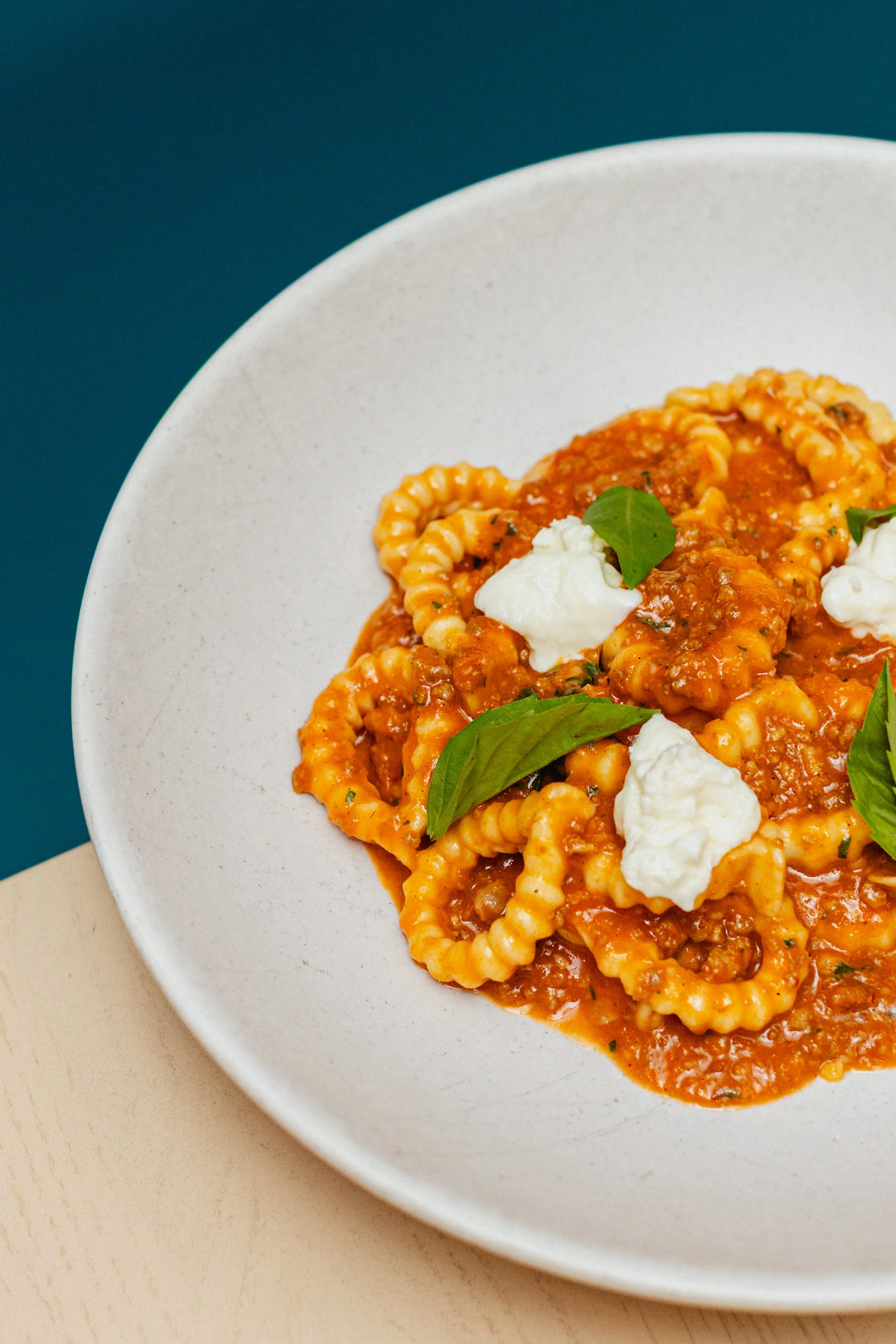 Close-up of a bowl of pasta with tomato sauce, garnished with basil leaves and dollops of white cheese, on a beige surface with a teal background.