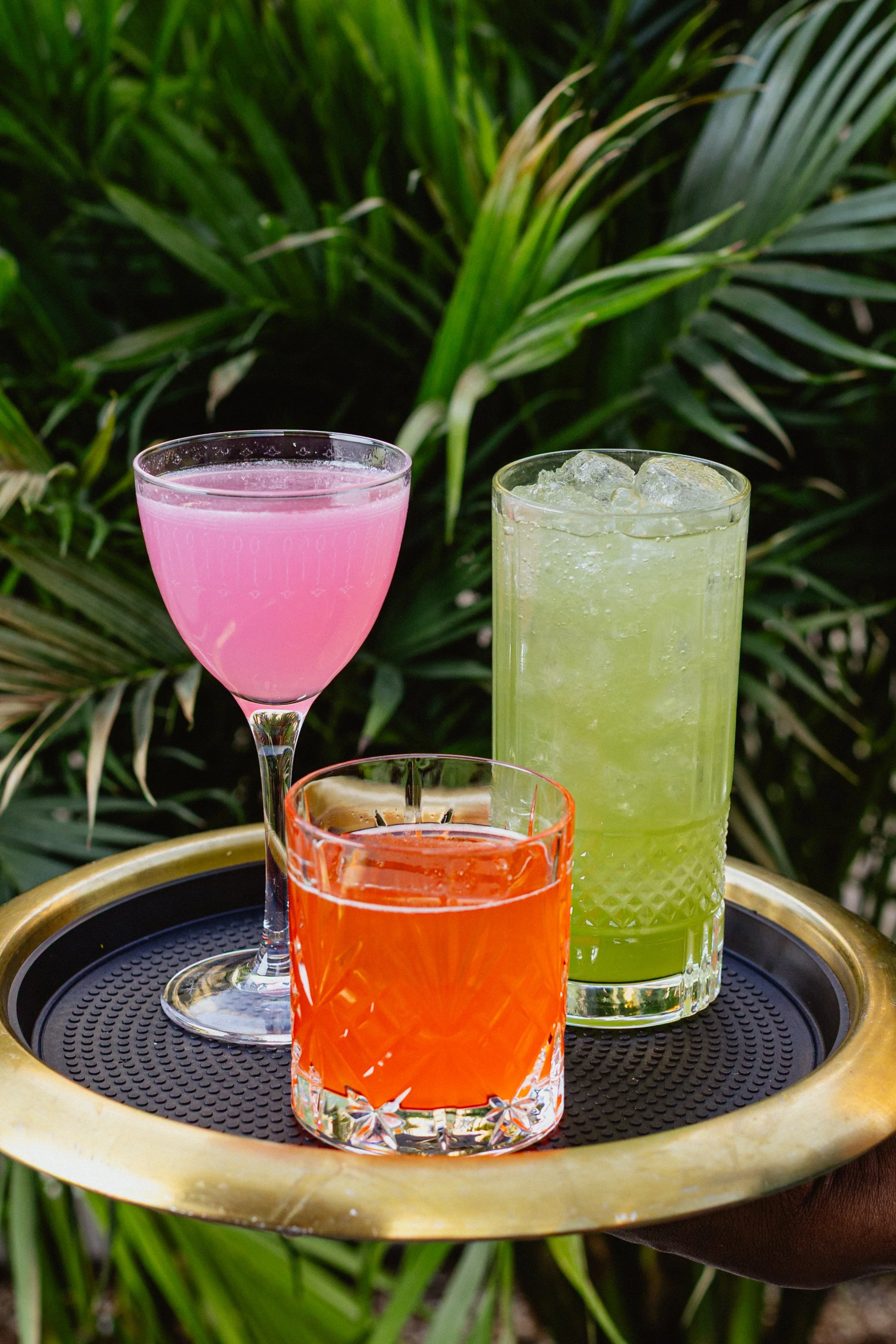 Four colorful drinks in different glasses on a round tray, placed outdoors with green plants in the background.