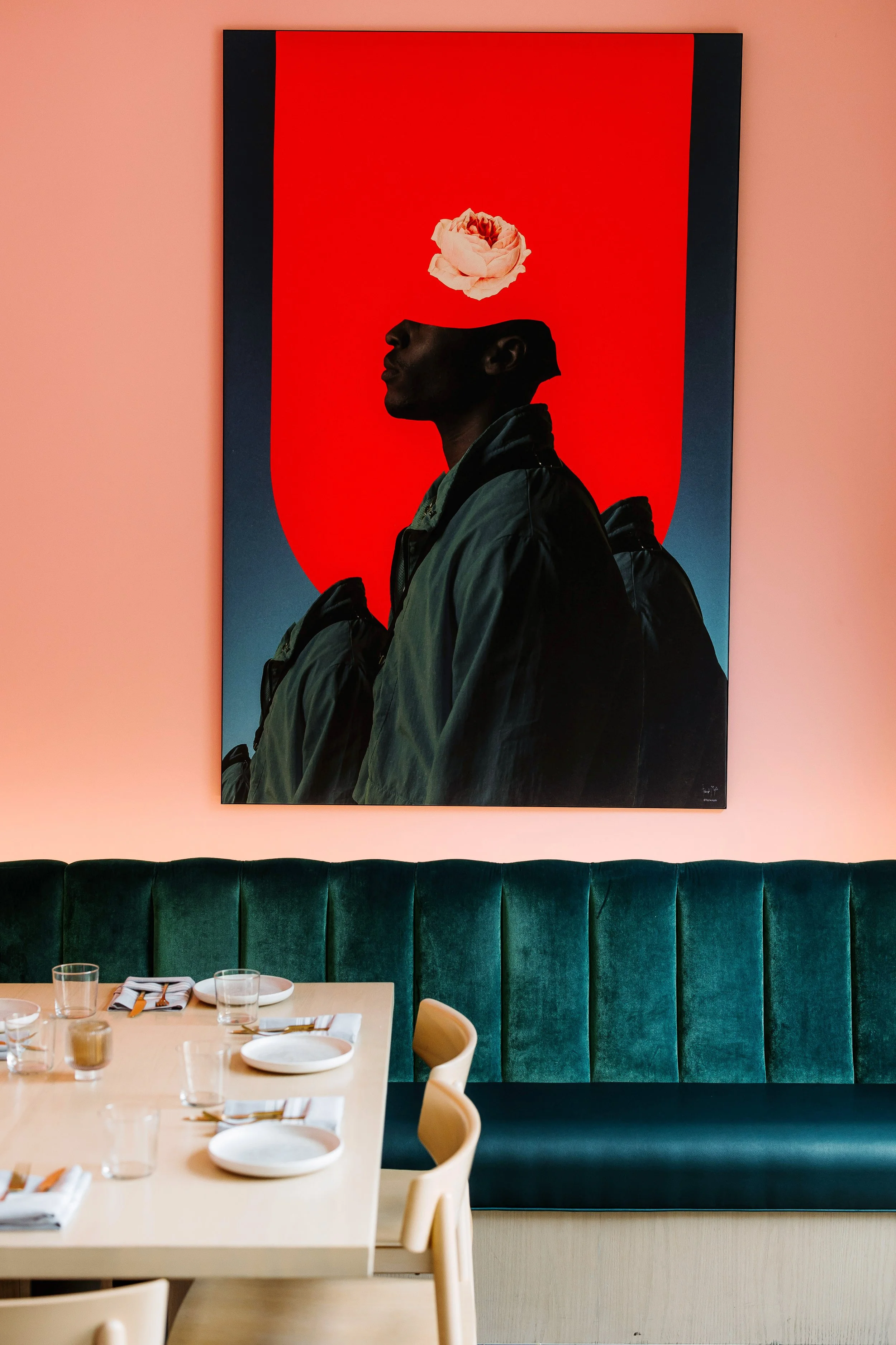 Interior of a restaurant with a green velvet bench along the wall, a table set with plates and glasses, and a large artwork of a silhouetted person with a pink flower above their head on a red background.