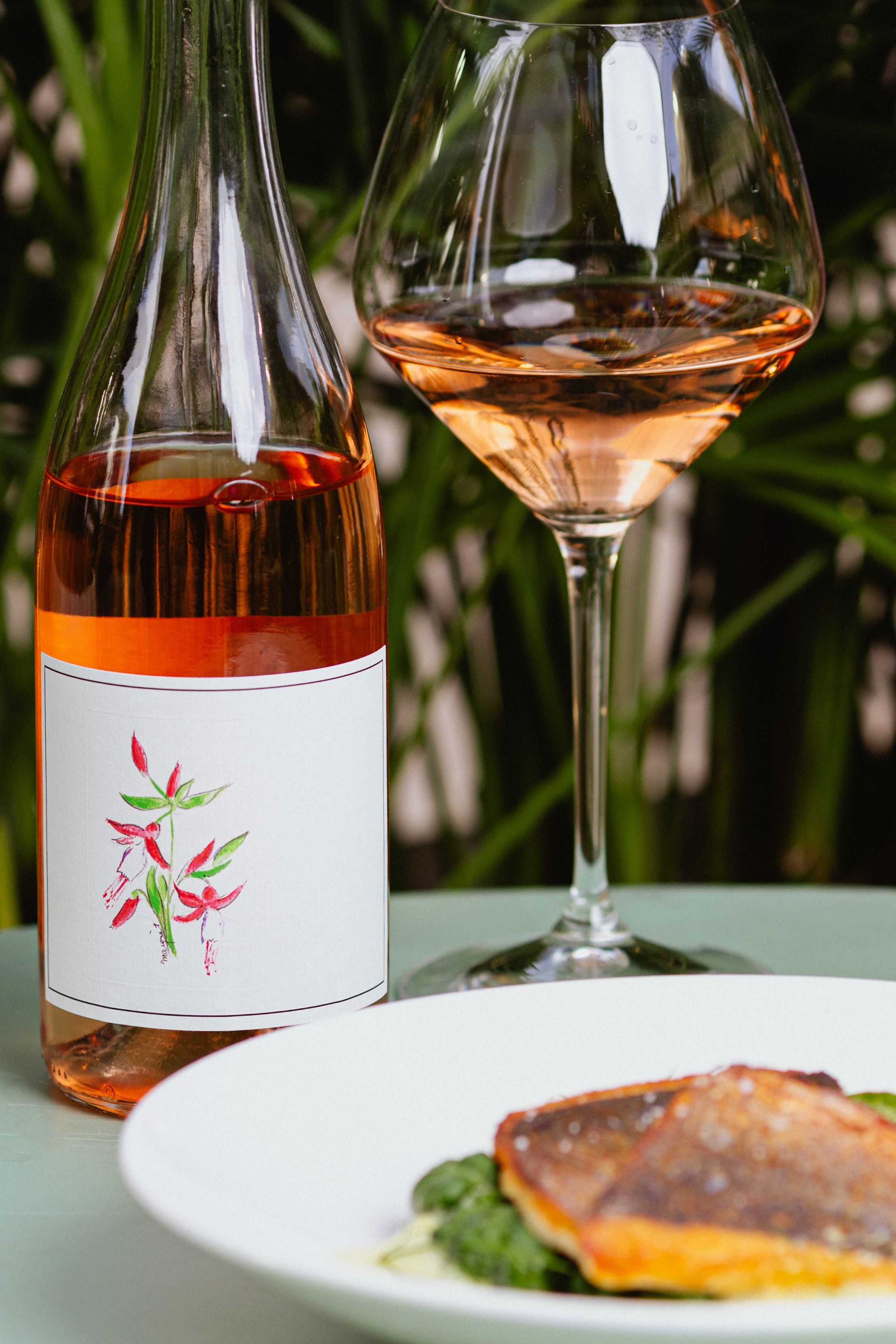 A bottle of rosé wine and a glass of rosé wine on a table with a plate of food in the foreground. The background has greenery.