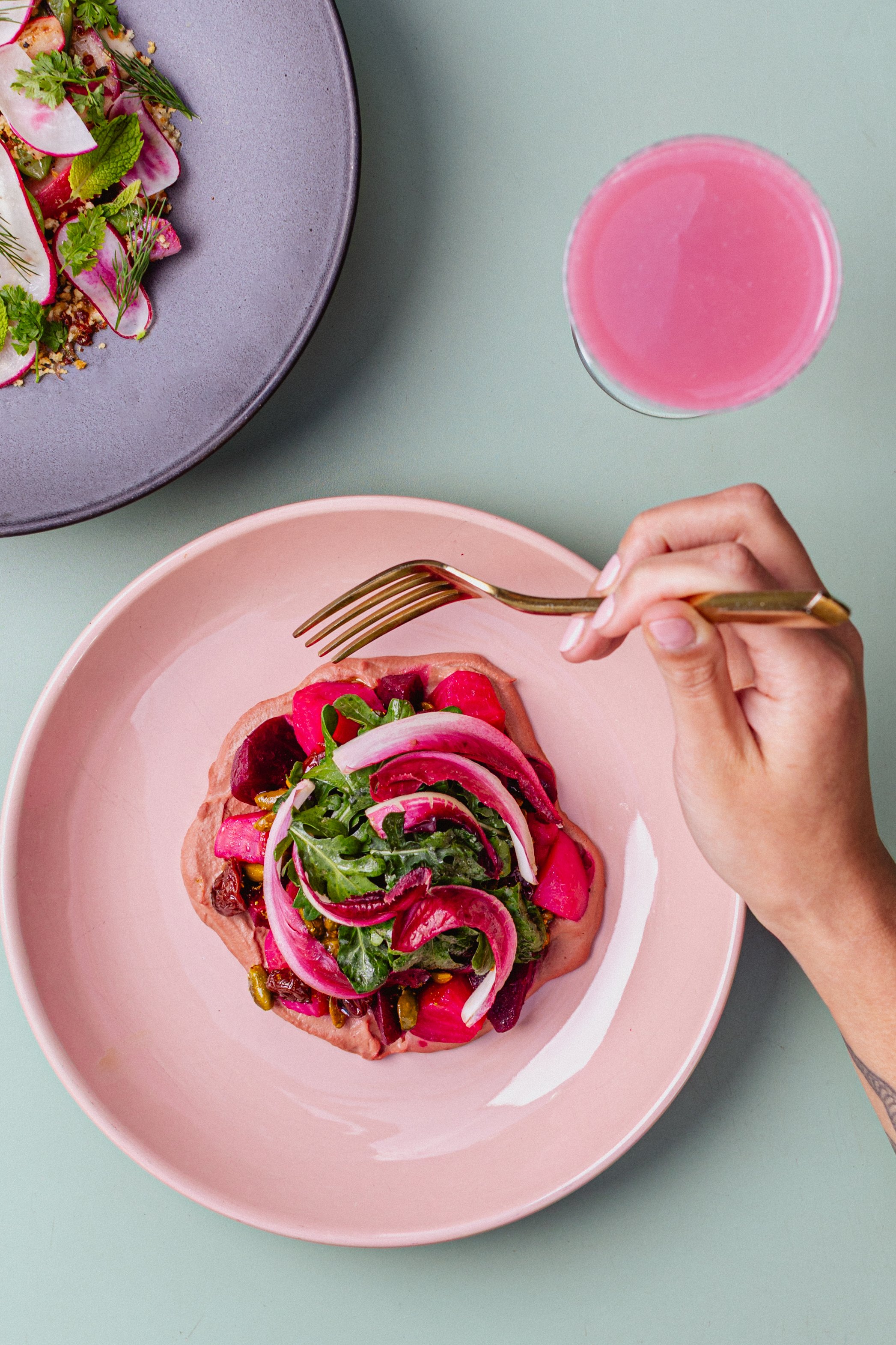 Fresh salad with radishes, greens, and sliced vegetables on a pink plate, a pink beverage in a glass, and a gray plate with salad in the background on a green surface.