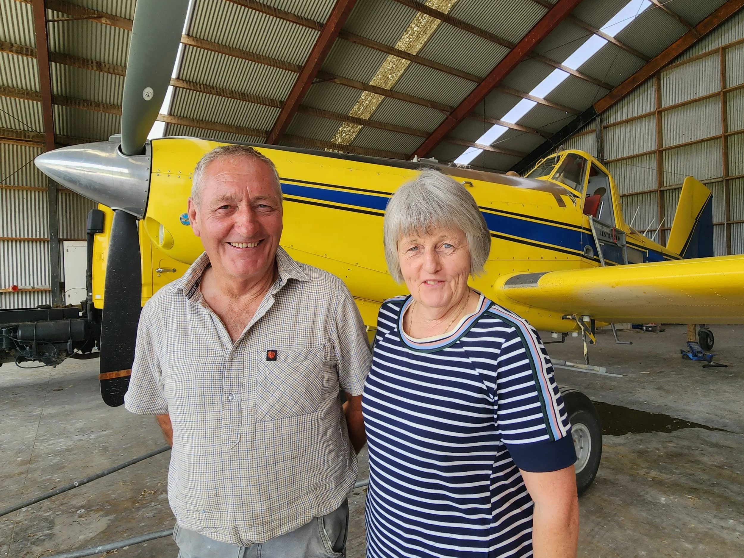 An older man and woman standing inside an airplane hangar with a yellow and blue aircraft in the background.