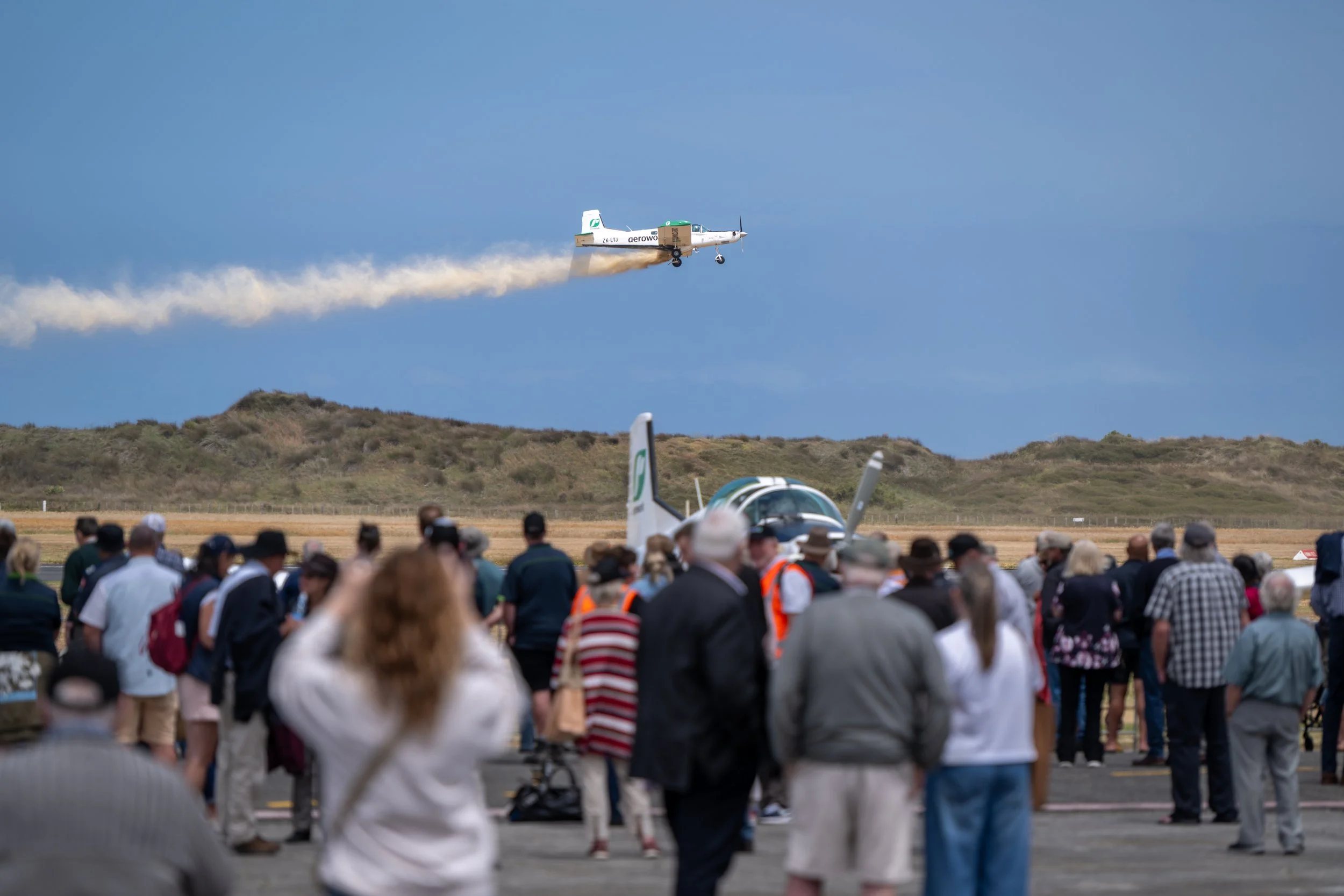 A crowd of people watching an airshow, with a small aircraft flying above and emitting smoke in the sky, and another aircraft parked on the ground.