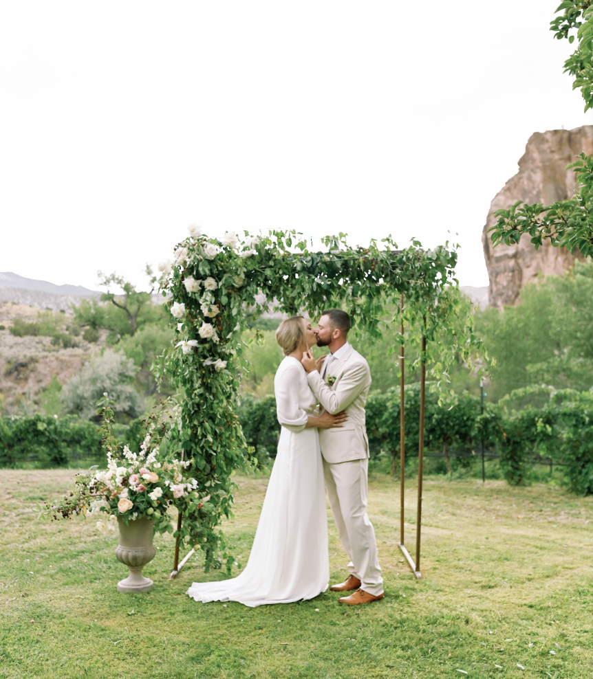 A bride and groom sharing a kiss under a decorated wedding arch in an outdoor, lush green setting.