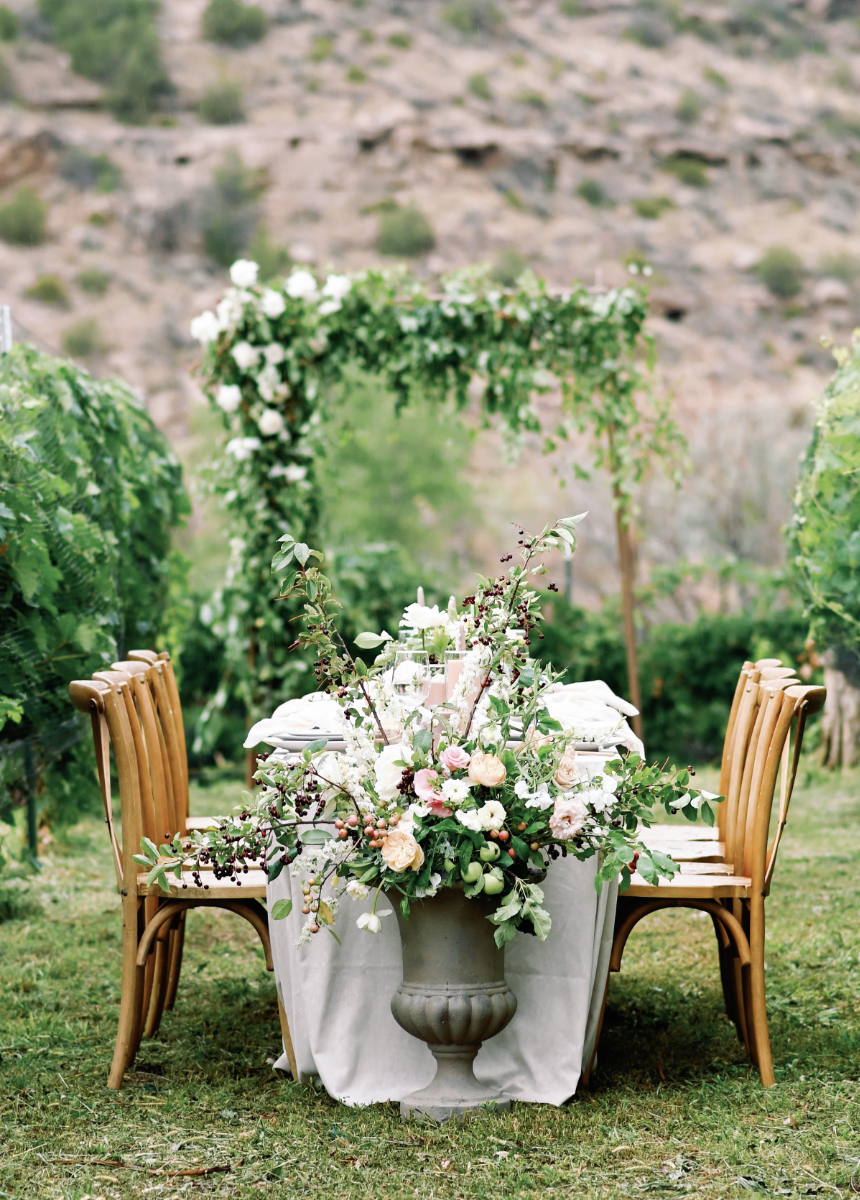 An outdoor dining setup for a wedding featuring a white tablecloth, wooden chairs, and a large floral centerpiece with white, pink, and green flowers, set in a garden area with greenery and mountain backdrop.