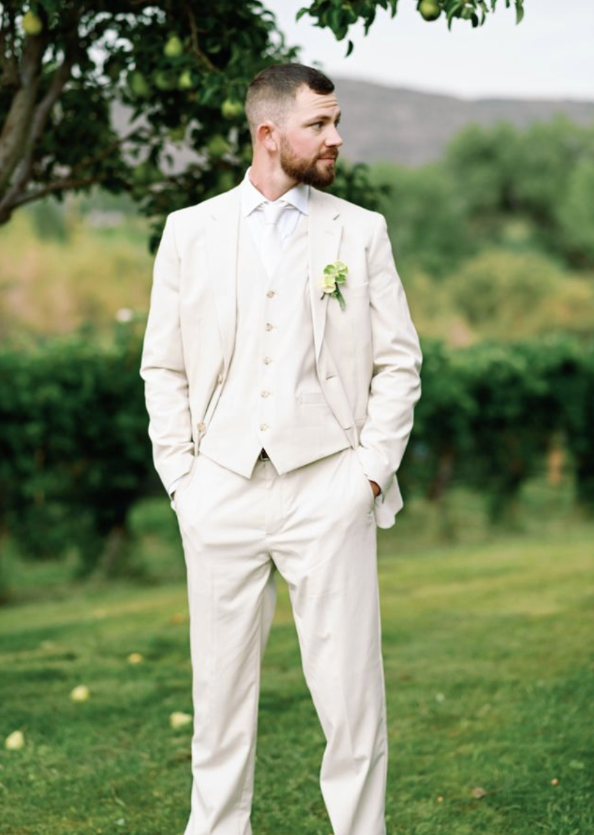 A groom in a white suit standing outdoors with hands in pockets, on a grassy area with trees and mountains in the background.