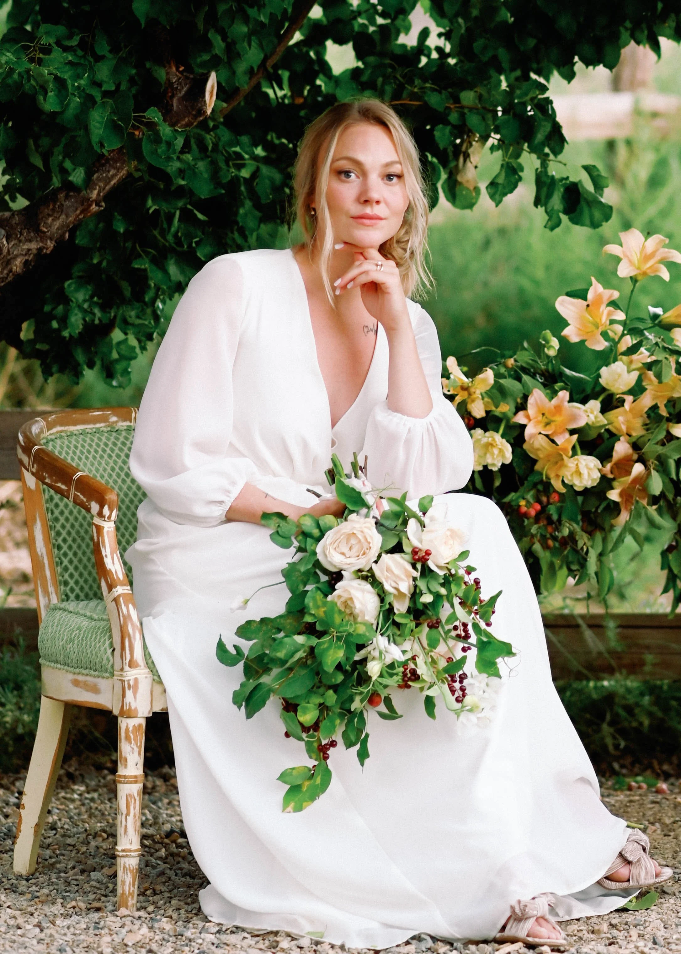 A woman dressed in a white gown sitting on a vintage chair outdoors, holding a bouquet of white and blush-colored flowers and greenery, with trees and flowers in the background.