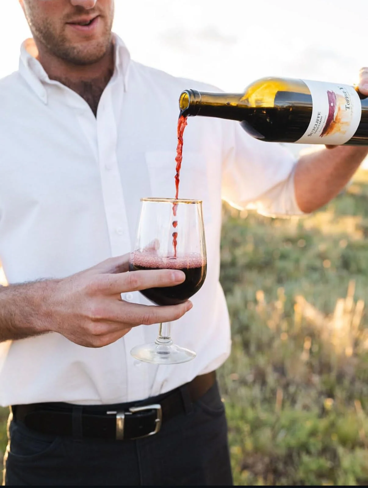 A man in a white shirt pours red wine from a bottle into a glass outdoors.