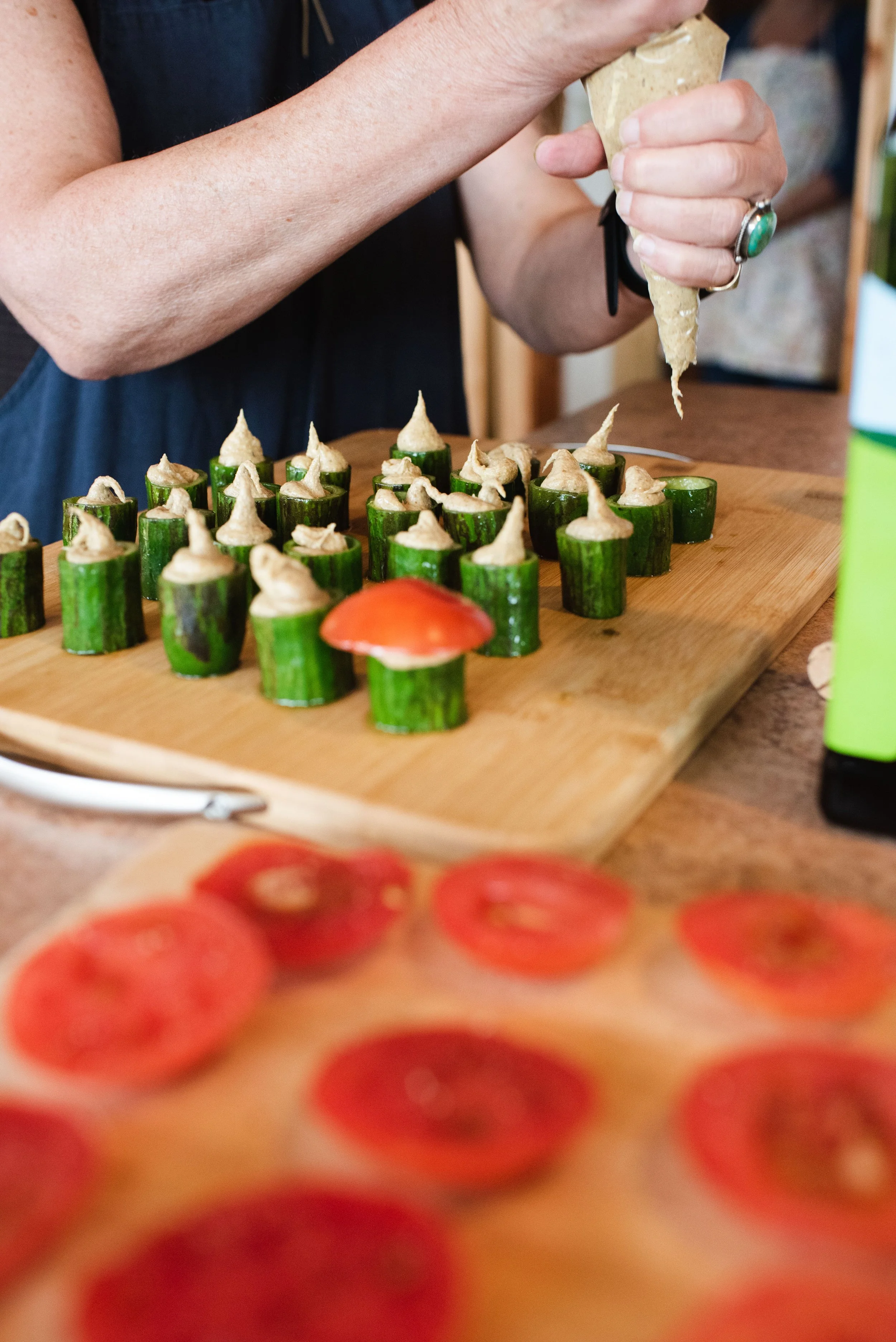 A chef is filling cucumber cups with a creamy spread at a wooden cutting board, with sliced tomatoes and a green bottle nearby.