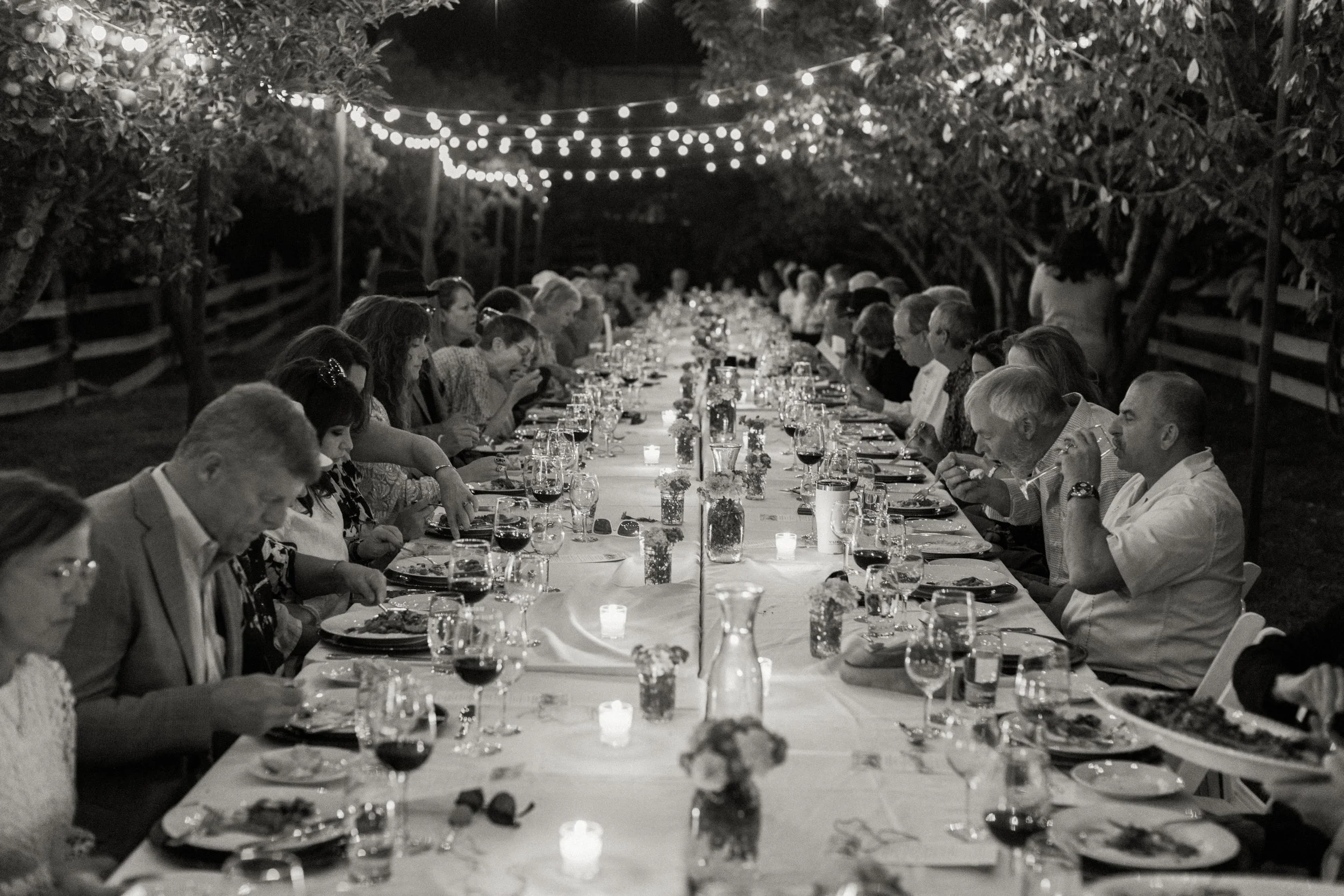 A long outdoor dinner table at night, decorated with flowers and candles, with many people seated on both sides, illuminated by string lights overhead.
