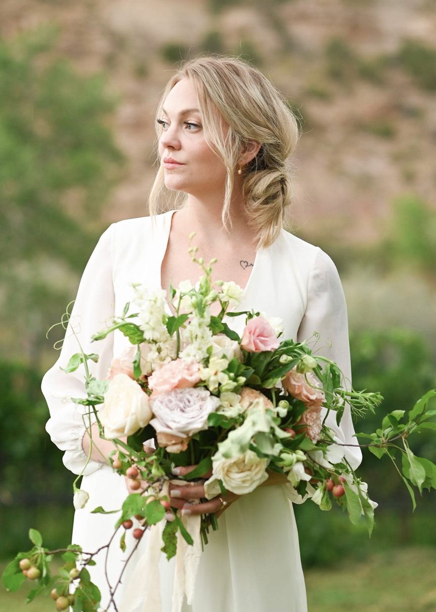 A bride in a white dress holding a large bouquet of flowers, standing outdoors with a background of greenery and a rocky hillside.