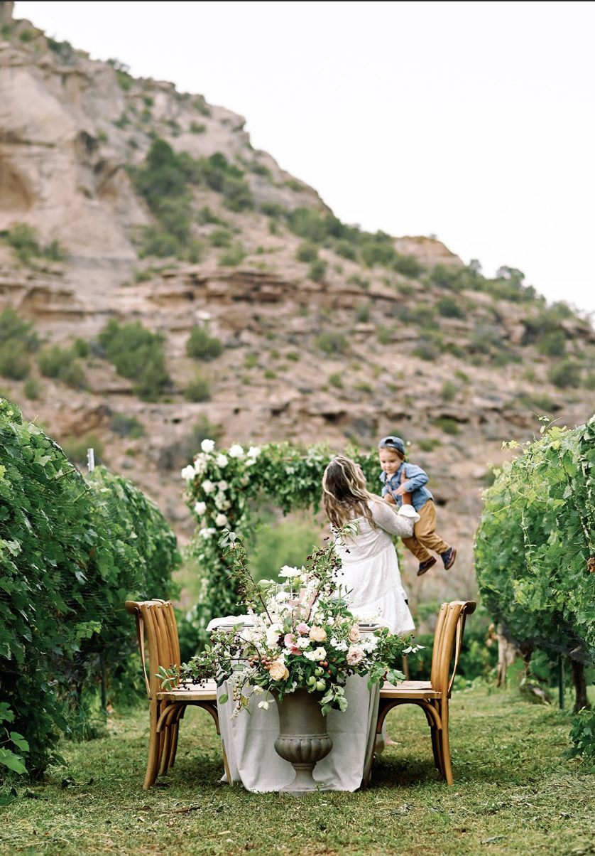 A woman at an outdoor wedding holding a young child in the vineyard next to a decorated table with a floral centerpiece, set outdoors near a mountain with lush green bushes and grapevines.