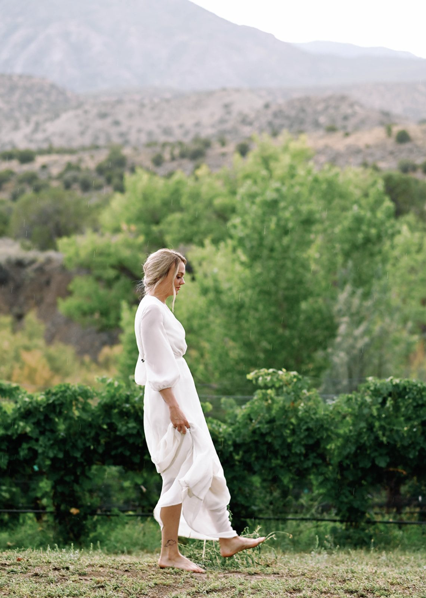 A woman in a white dress walking barefoot outdoors with lush green trees and mountains in the background.