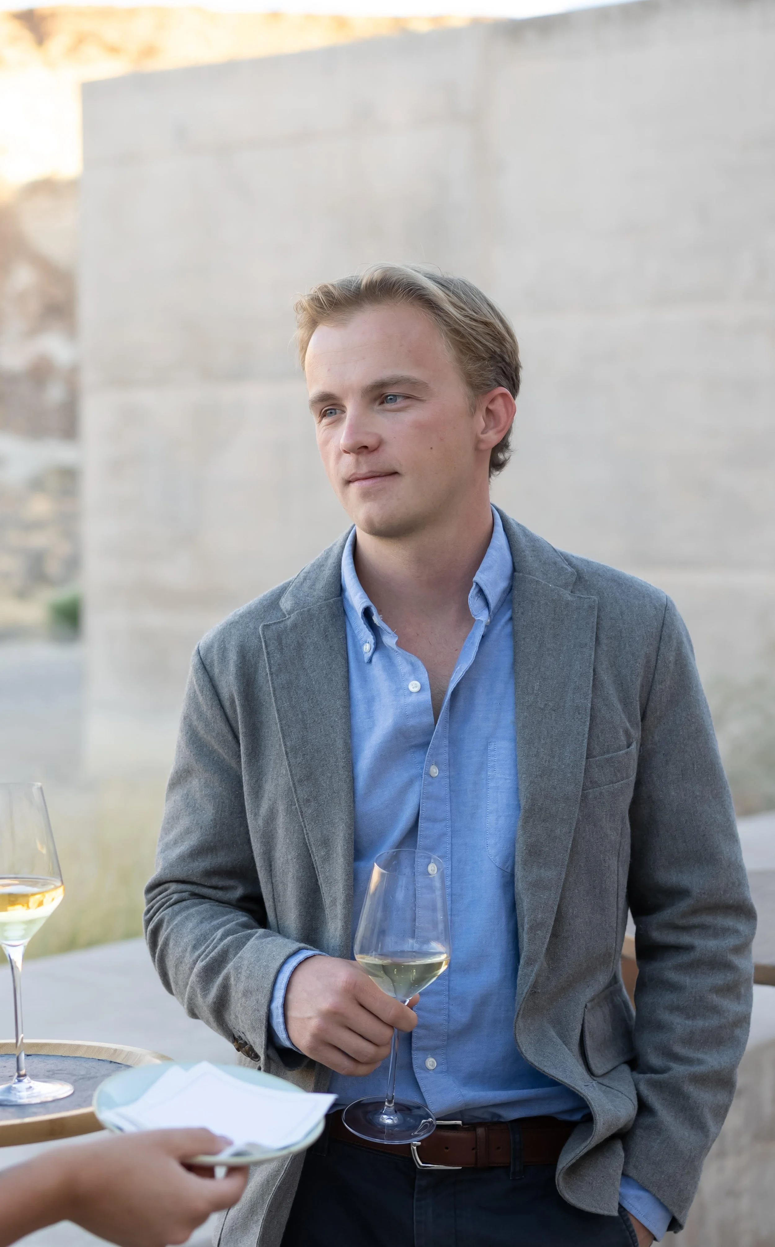 A man with blonde hair in a gray jacket is holding a glass of white wine in a desert resort setting.