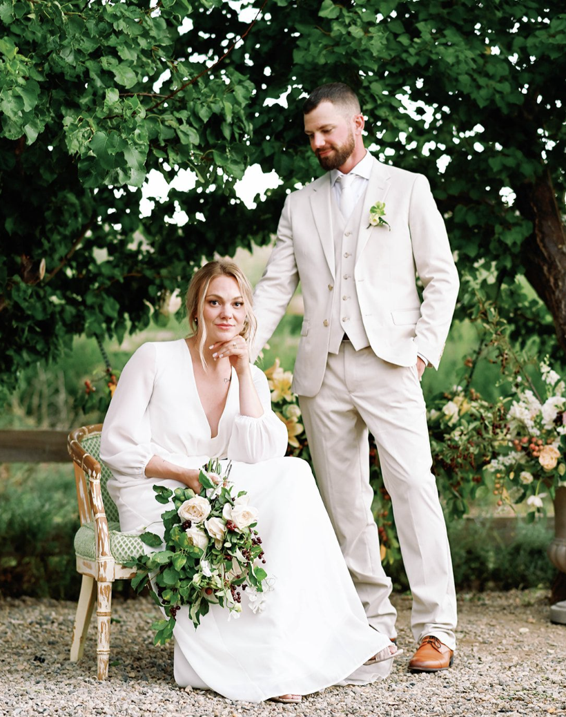 A bride sitting on a vintage chair with a bouquet, and a groom standing beside her with his hand on her shoulder, outdoors under a green leafy tree, in wedding attire.