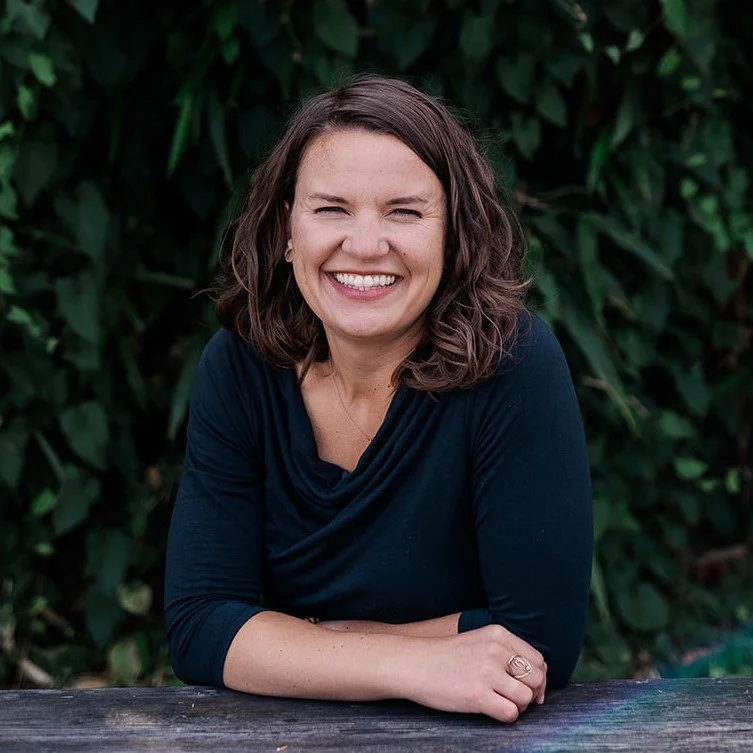 A brown-haired woman who is a pelvic floor physical therapist is smiling, wearing a ring with female anatomy depicted. She is sitting in front of greenery in Tacoma, Washington with a friendly, passionate smile that is welcoming and sensitive.