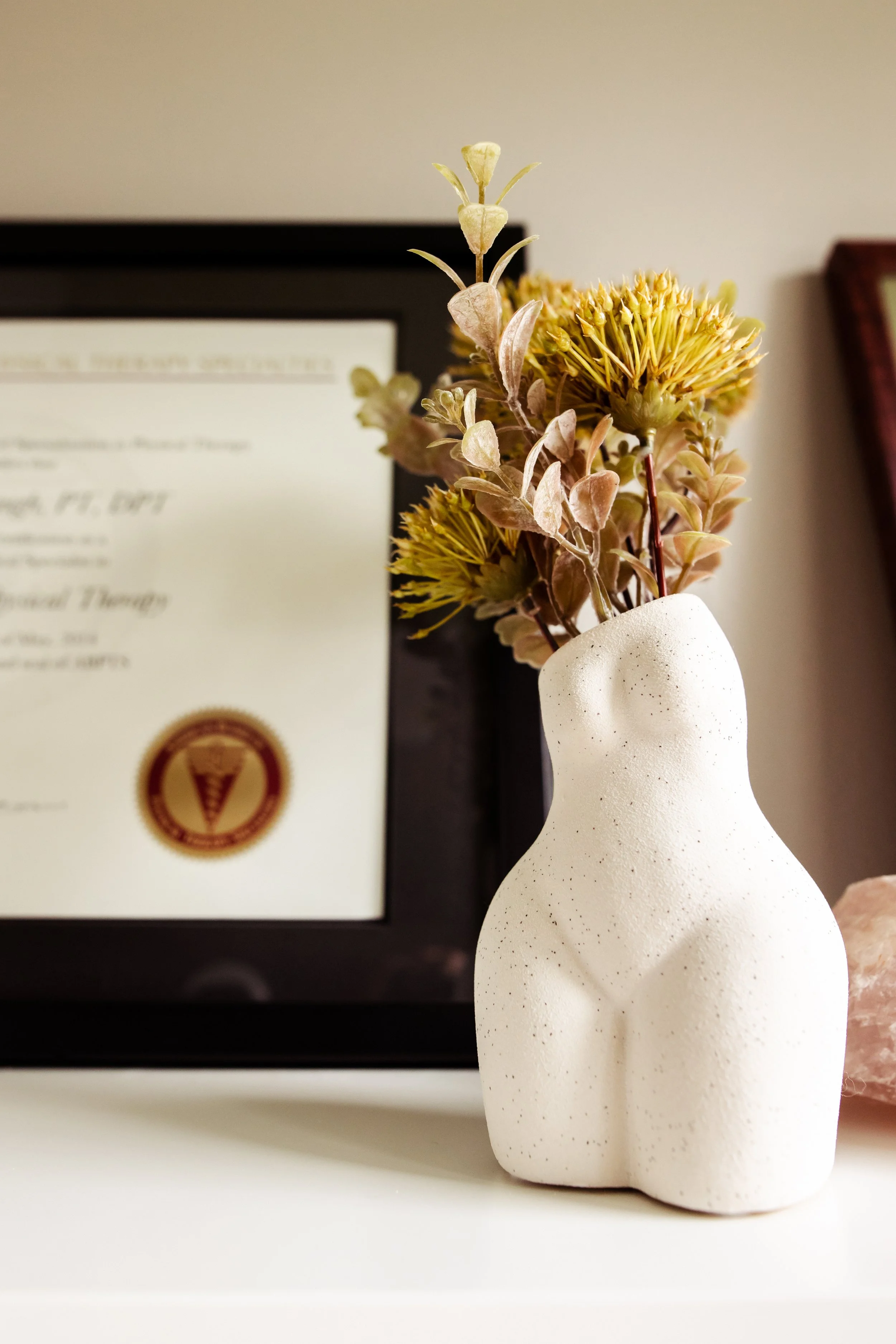 A small white ceramic vase with a feminine figure holds dried yellow and pink flowers. In the background, a framed Board-certification in Women's Health certificate is partially visible on the wall.