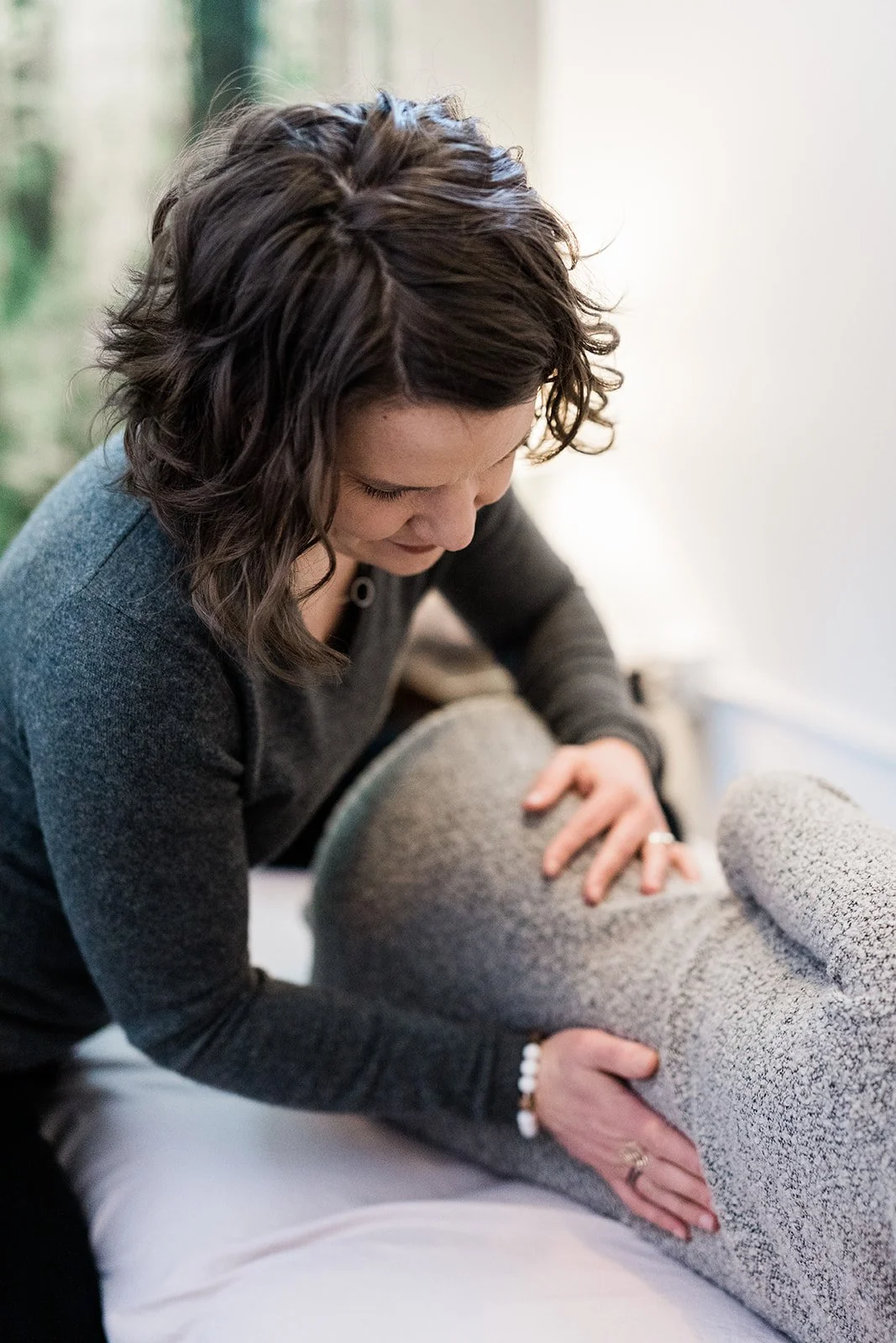 A woman who is a pelvic floor physical therapist performing a gentle spine and hip adjustment on a pregnant person lying on a table in a cozy room.