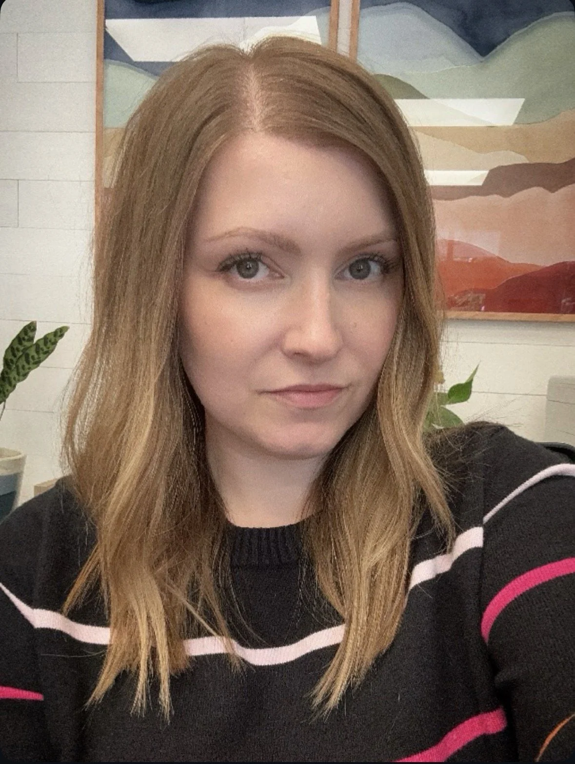 A woman with long, wavy hair is wearing a black sweater with stripes to represent the Pride flag. She is sitting behind the front desk with colorful abstract art and potted plants behind her, welcoming patients to the inclusive pelvic floor PT clinic