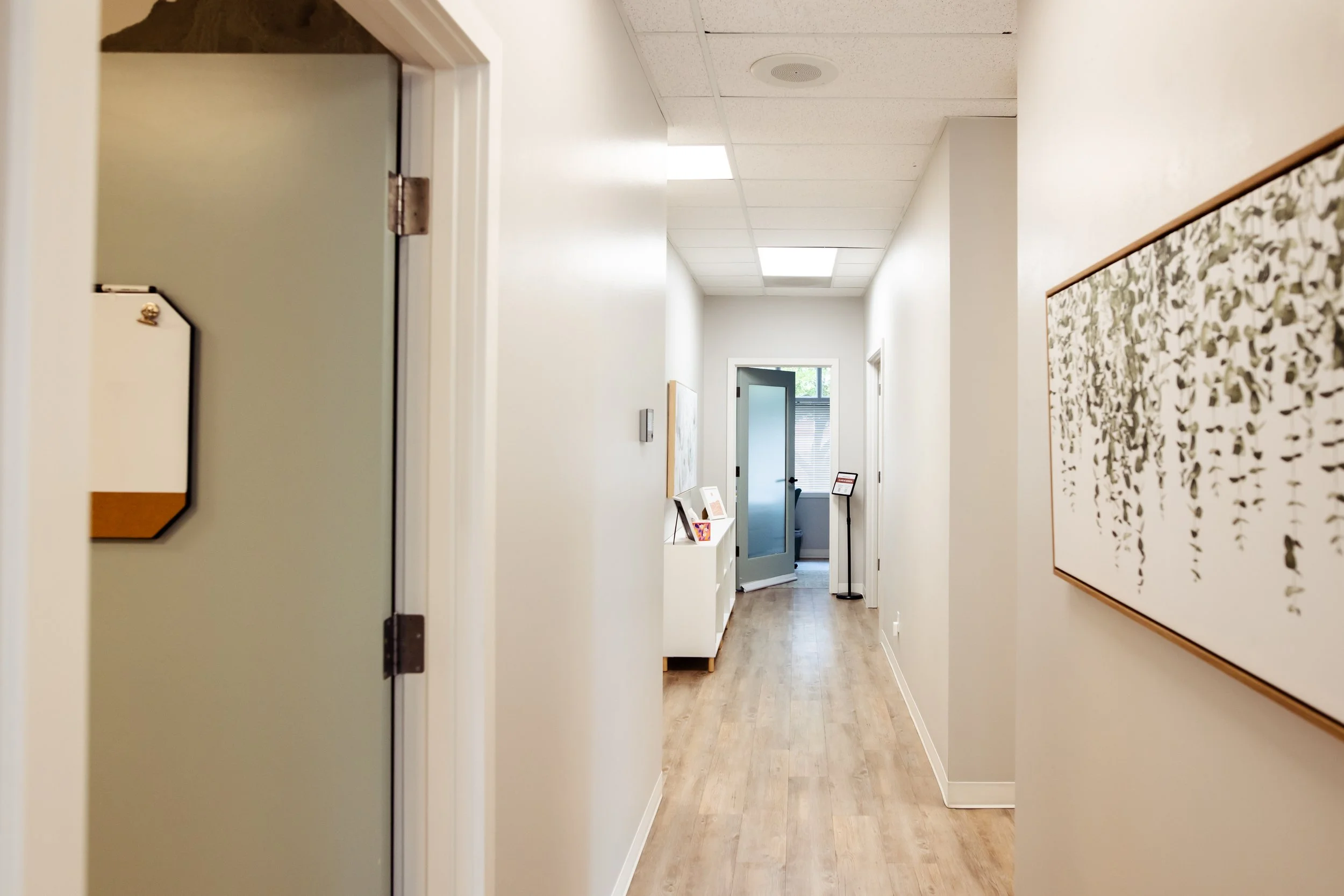 A welcoming, open indoor hallway with white walls, a wooden floor, and a drop ceiling with soft lights welcoming you into the inclusive healing space. Several doors and a large framed artwork are visible along the hallway.