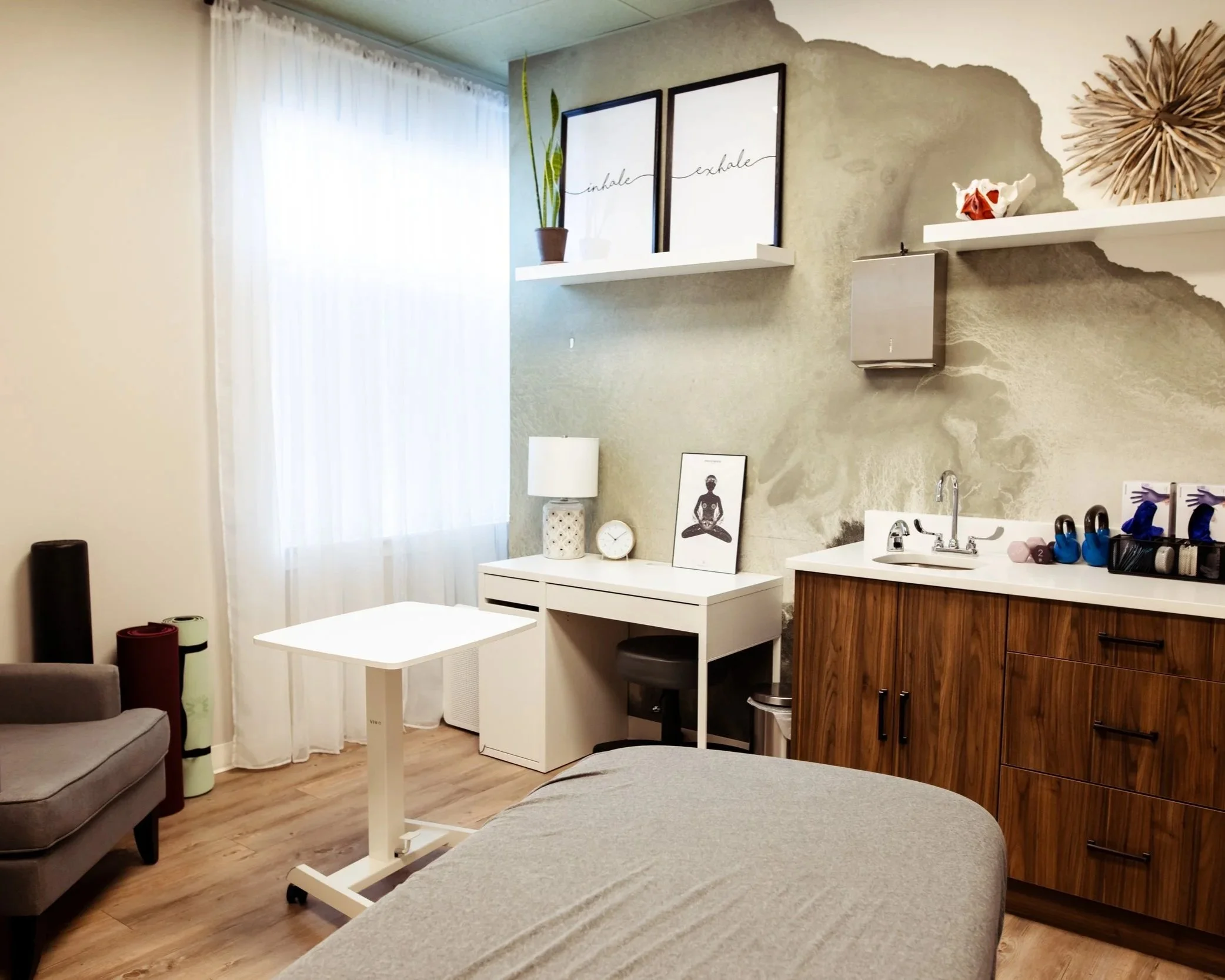A private treatment room with a massage table and cotton sheets. The wall behind features trauma-sensitive yoga-style art. The room is lit by natural light coming through sheer white curtains. To the right is a sink, a cabinet and various PT tools.