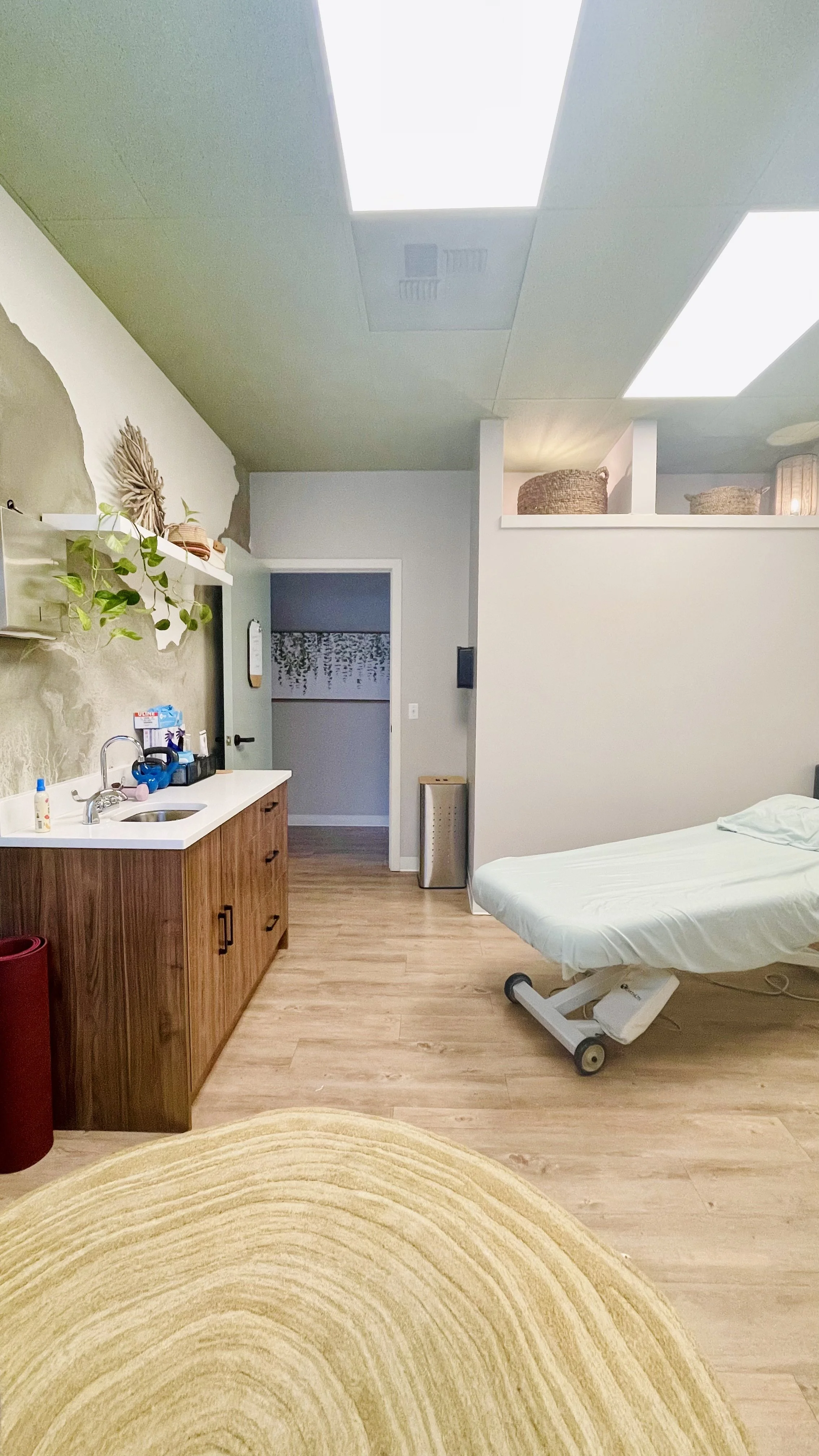 A private women's health PT treatment room with a massage table with soft sheets on the right, a wood cabinet with a sink on the left. The ceiling has green panels with soft lights, and a decorative wall on the left with plants and woven baskets.