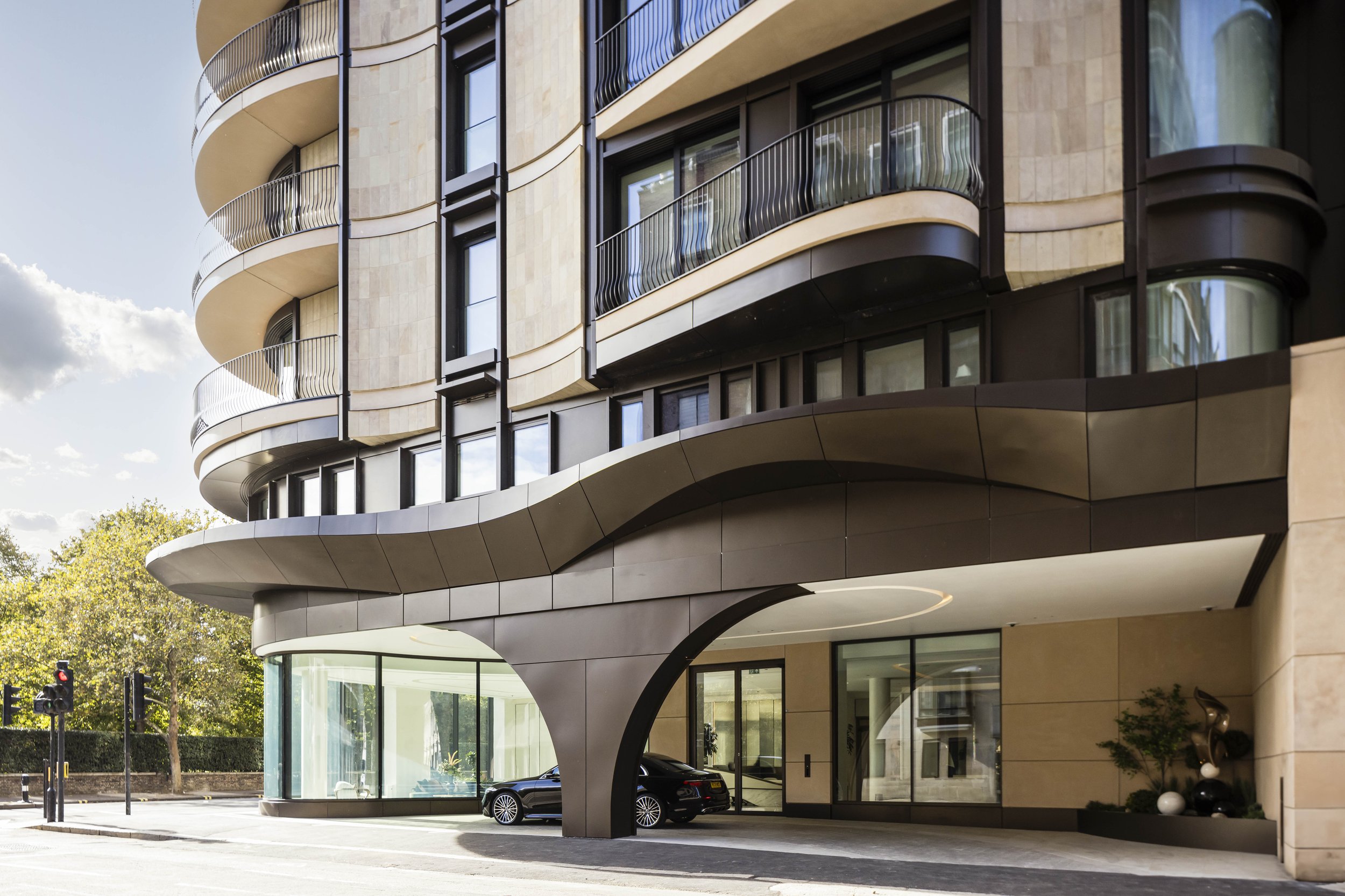 Modern multi-story building with curved balconies, large glass windows, and sleek architectural design. There are cars parked underneath and trees lining the street.