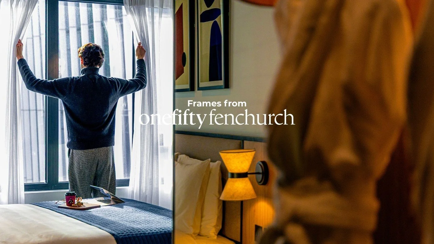 Person opening curtains in a brightly lit hotel room with a bed, artwork, and a wall lamp.